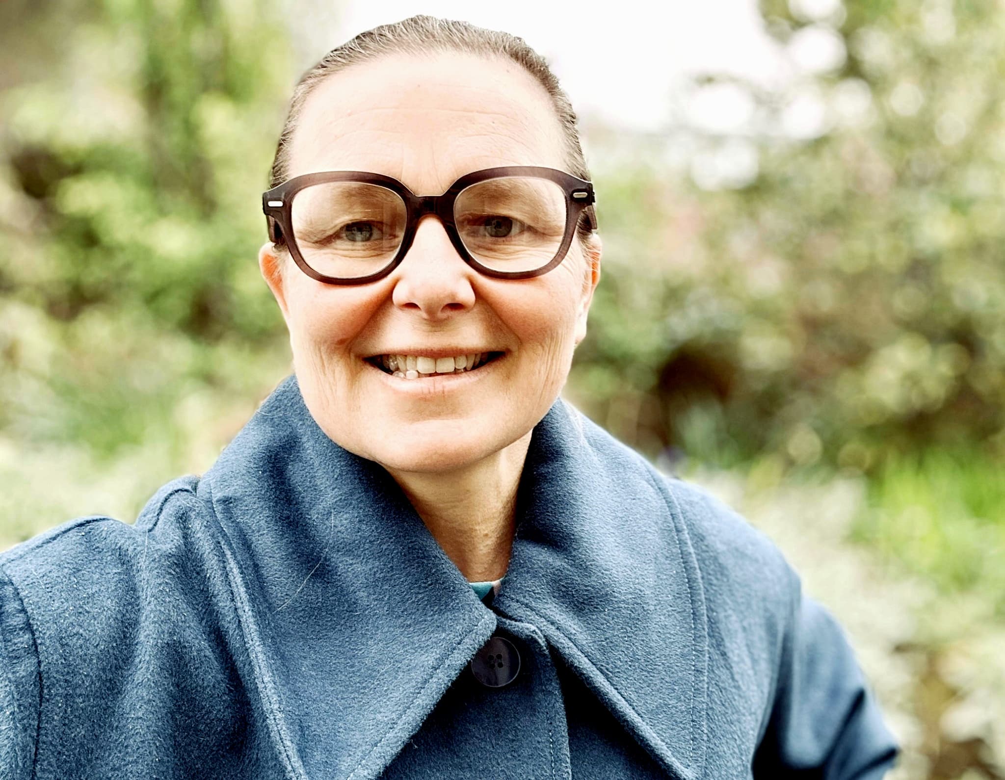 Smiling middle-aged woman with tortoiseshell glasses and slicked-back hair wearing a blue wool coat outdoors with blurred greenery behind her.