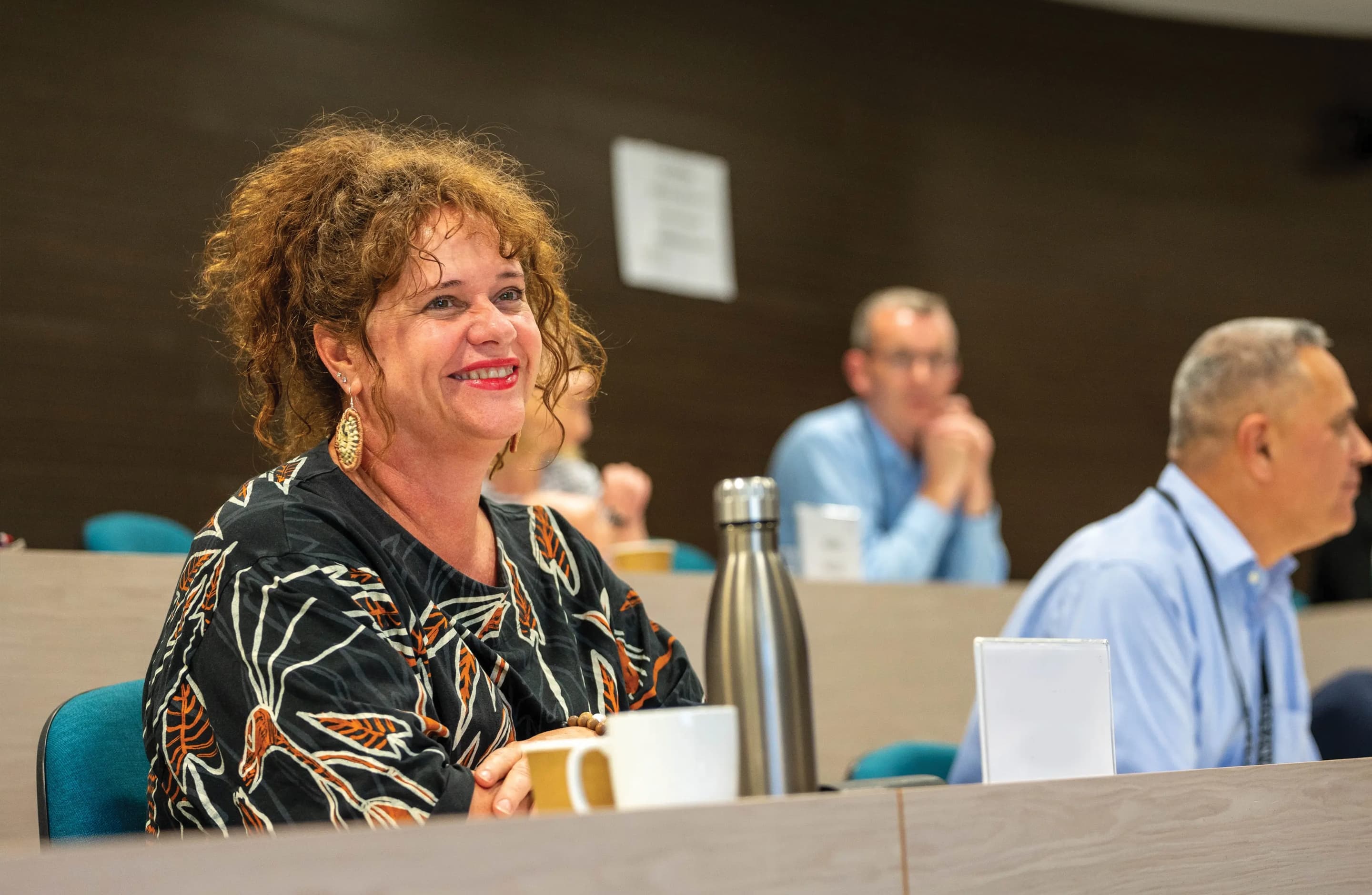 Woman with curly hair smiling at a conference table with a coffee mug and water bottle, two men seated in the background.