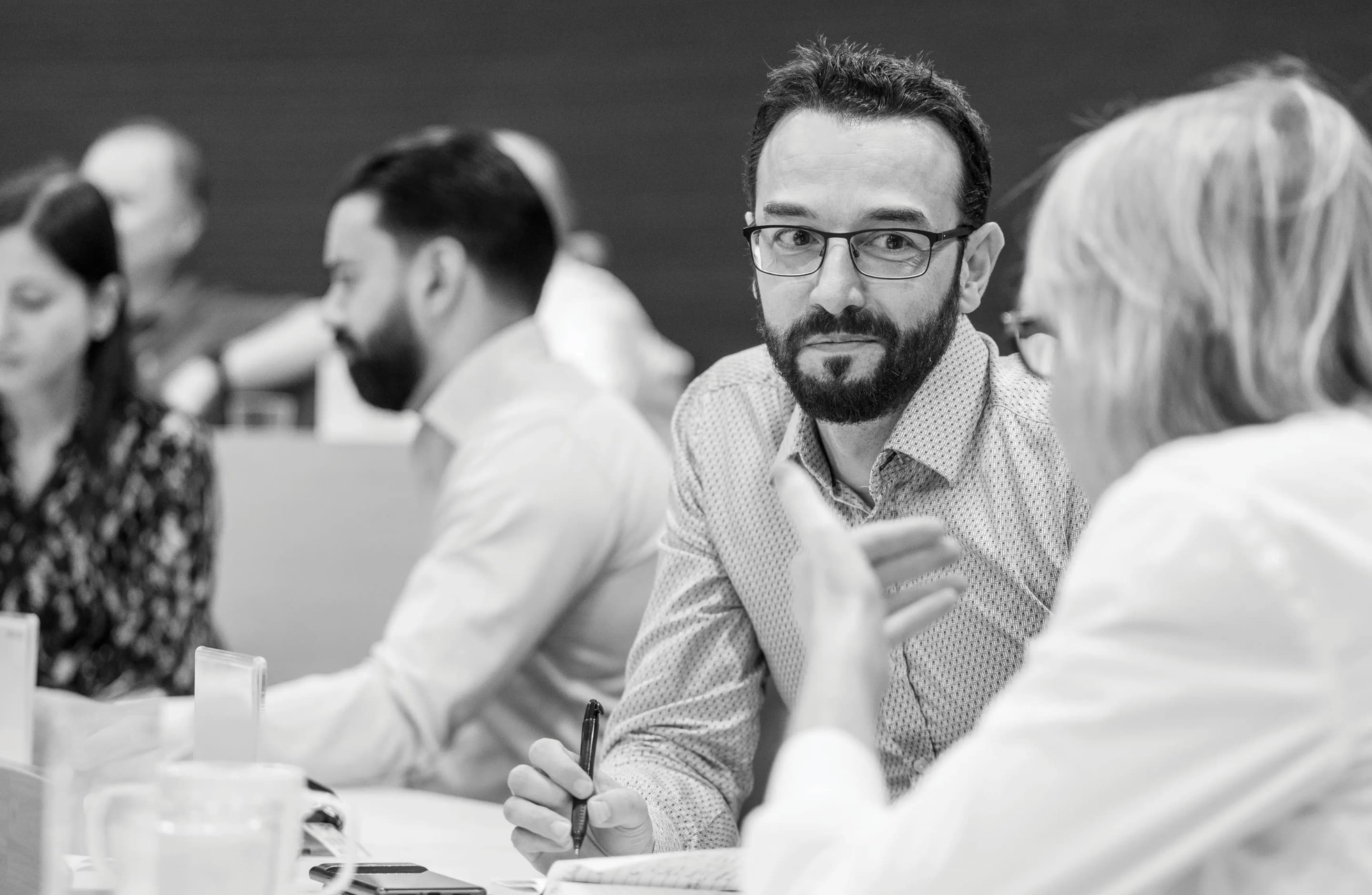 Bearded man with glasses holding a pen listens attentively during a business meeting or workshop, with other participants blurred in background.