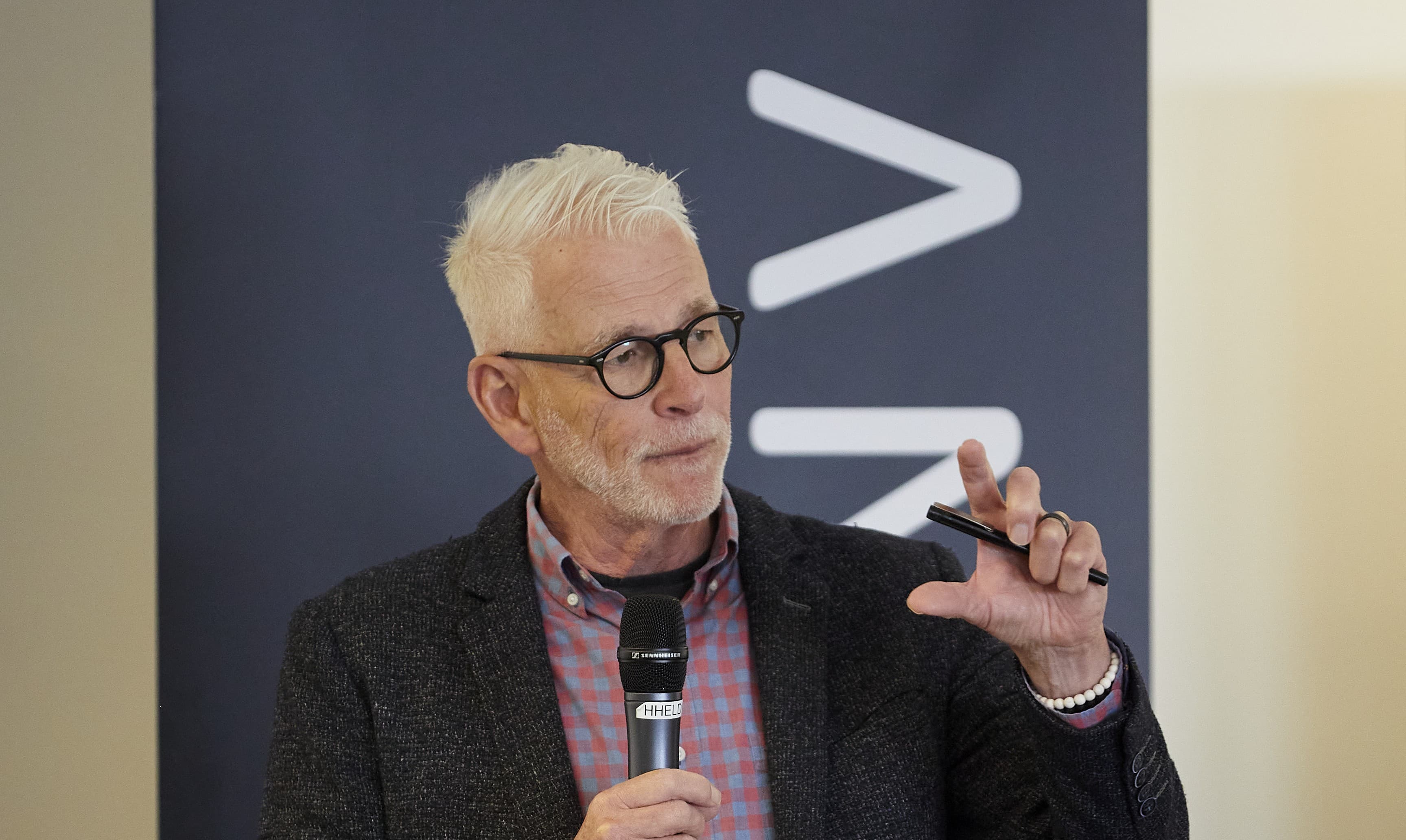 Elderly man with glasses speaking into a microphone, gesturing with a pen. Background features dark banner with white emblem.