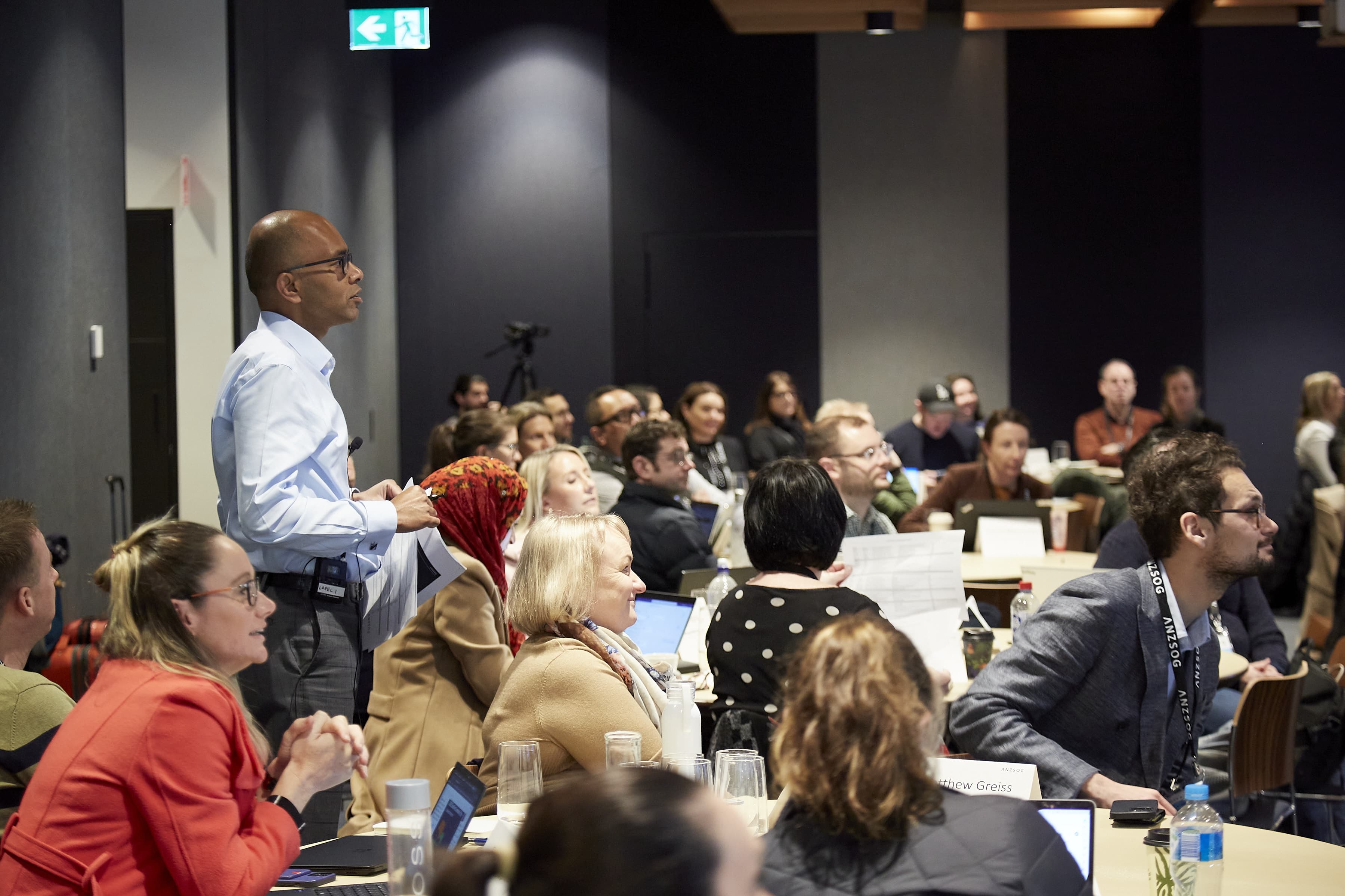 Attendees at a conference seated at tables with laptops listen as a man stands up and speaks to the room.