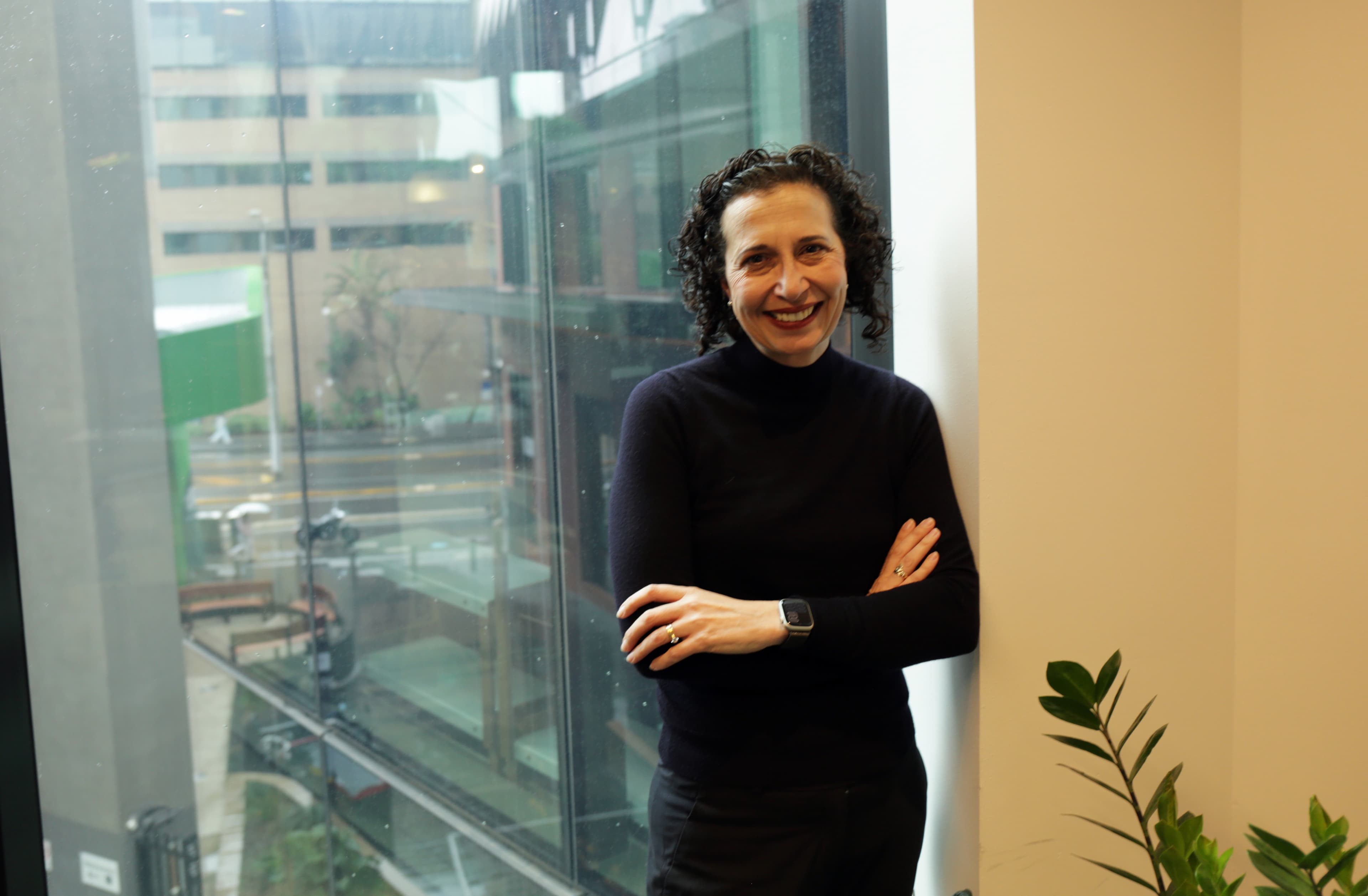 A smiling woman with curly dark hair in a black turtleneck stands arms crossed by a large office window overlooking a city courtyard.