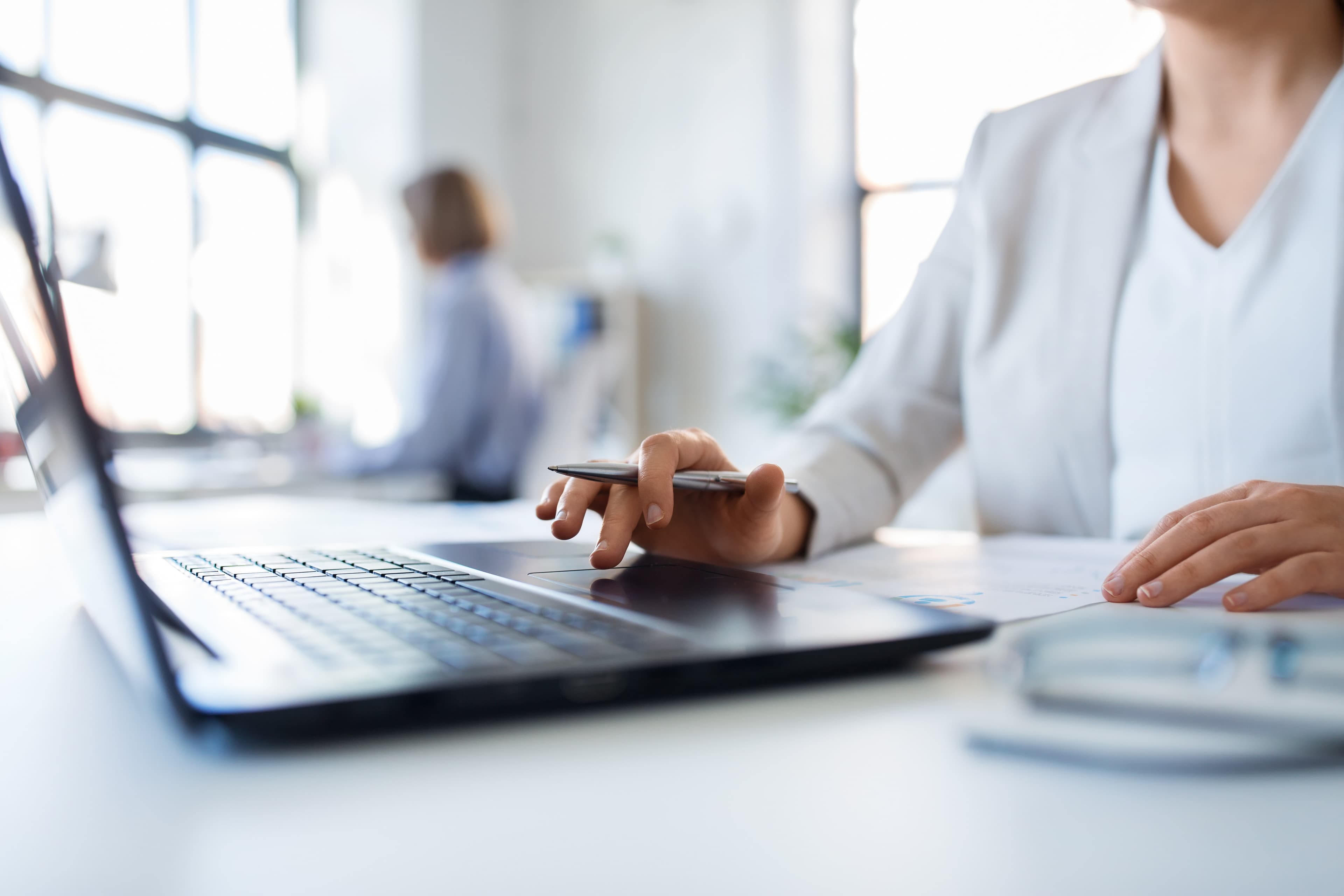 A person in a white outfit works on a laptop, holding a pen, in a bright office with large windows. Another person is blurred in the background.