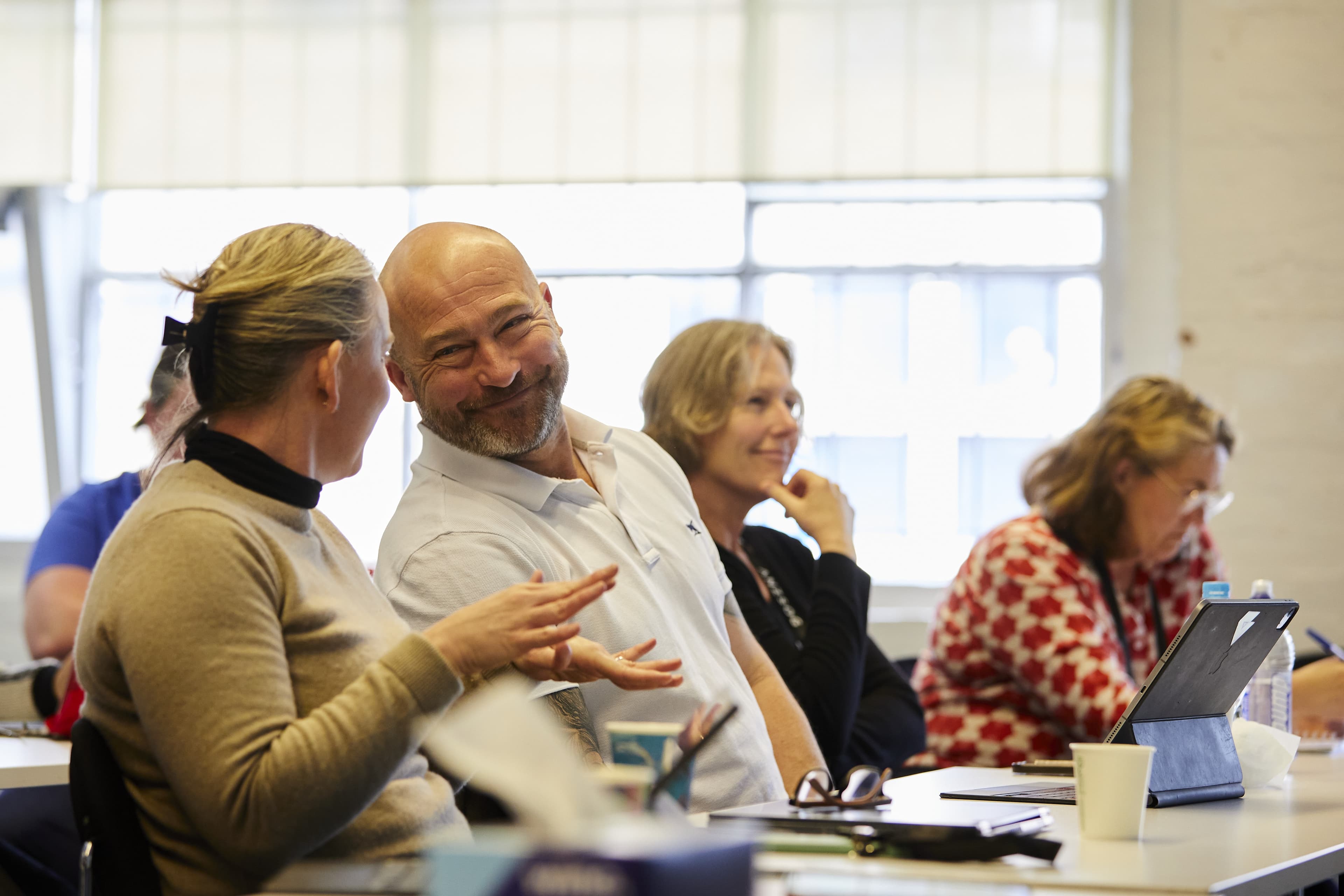 People seated in a bright classroom; smiling man leans toward a woman while others listen, write, and use a tablet.