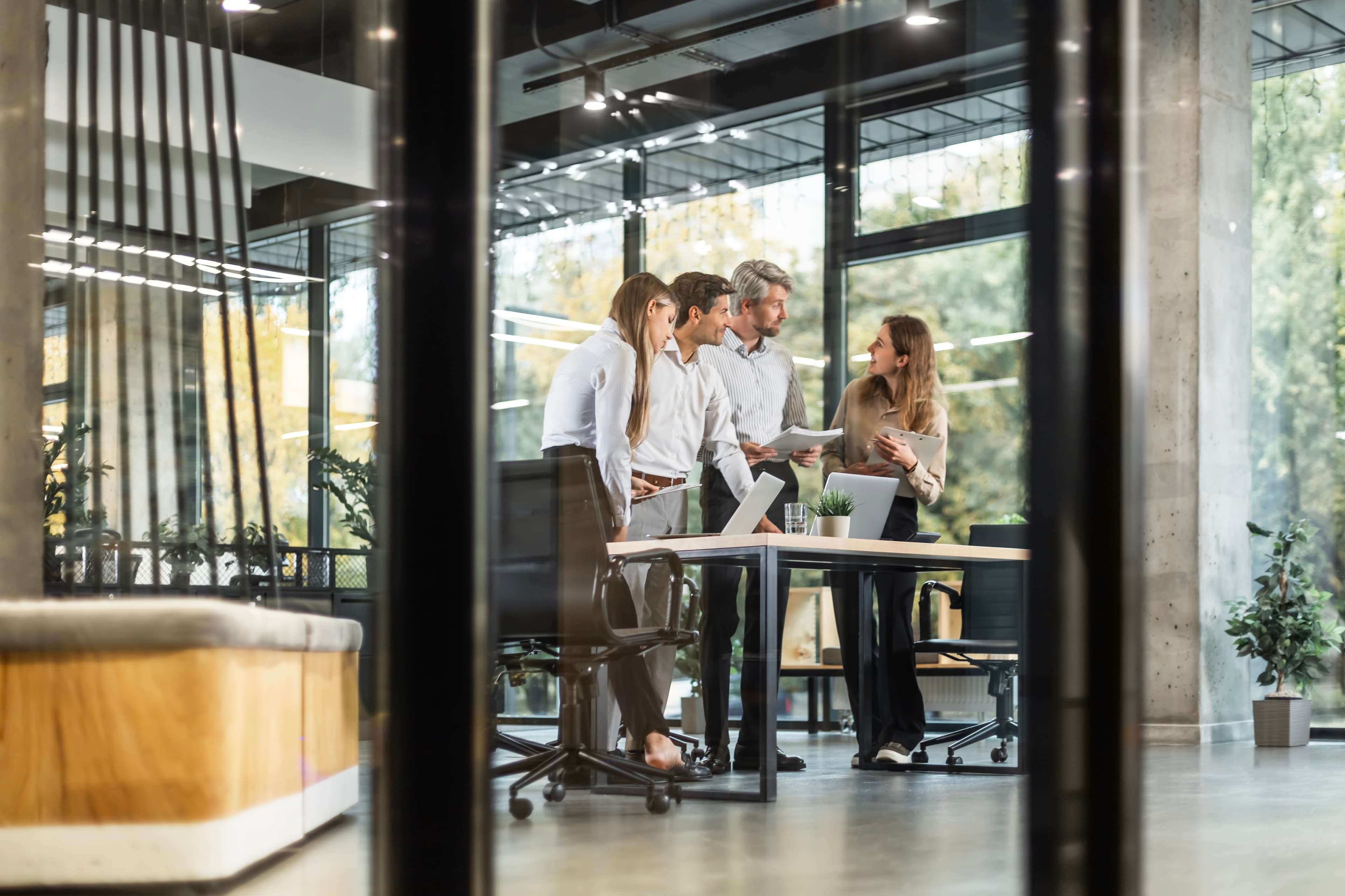 Four people in business attire stand around a table, discussing documents in a modern office with large windows and plants.