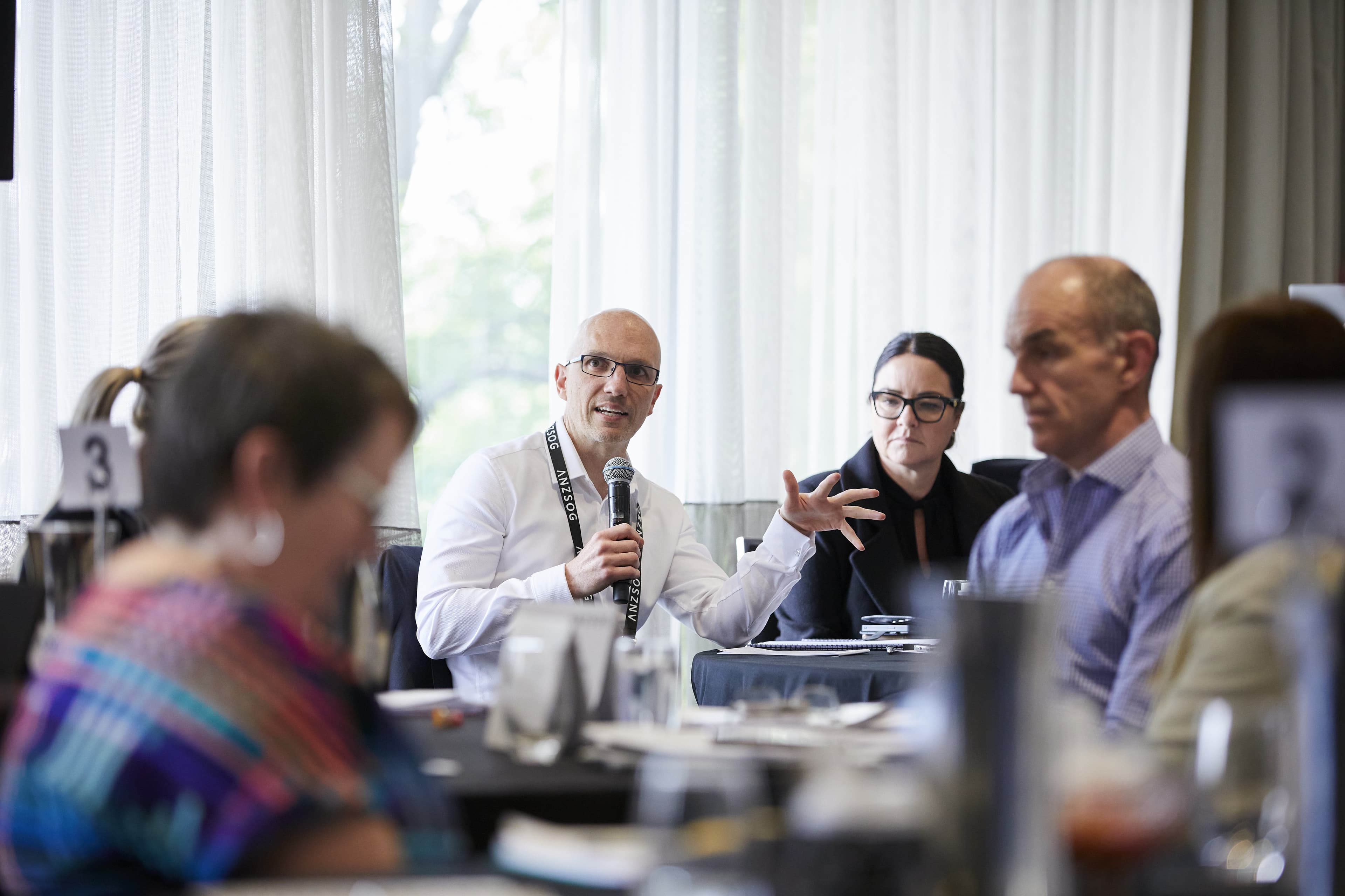 Man holding a microphone speaks while colleagues at a conference table listen attentively; a woman beside him gestures.