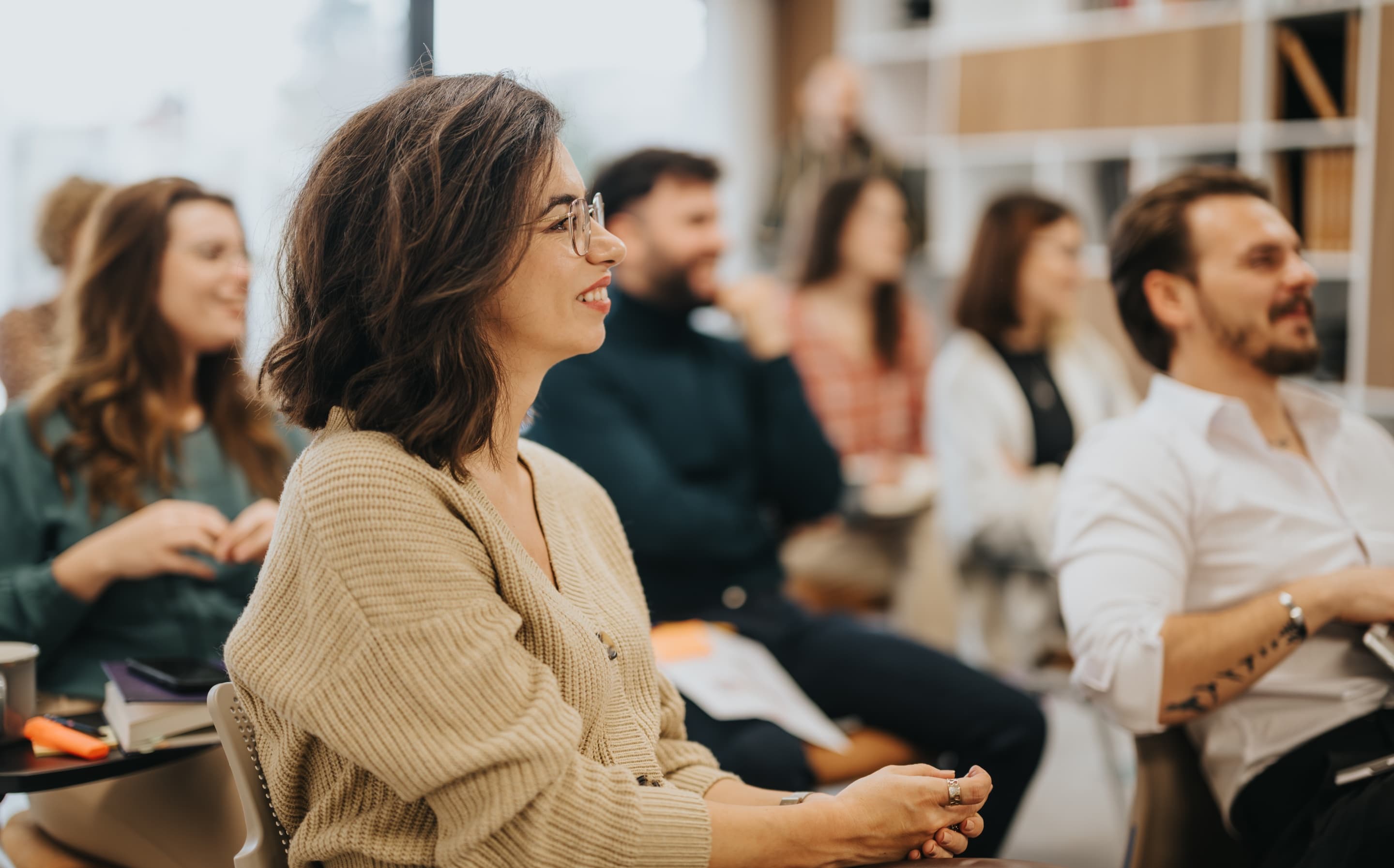 Smiling young woman with glasses in a beige sweater sitting among engaged, laughing attendees at an indoor seminar or workshop.