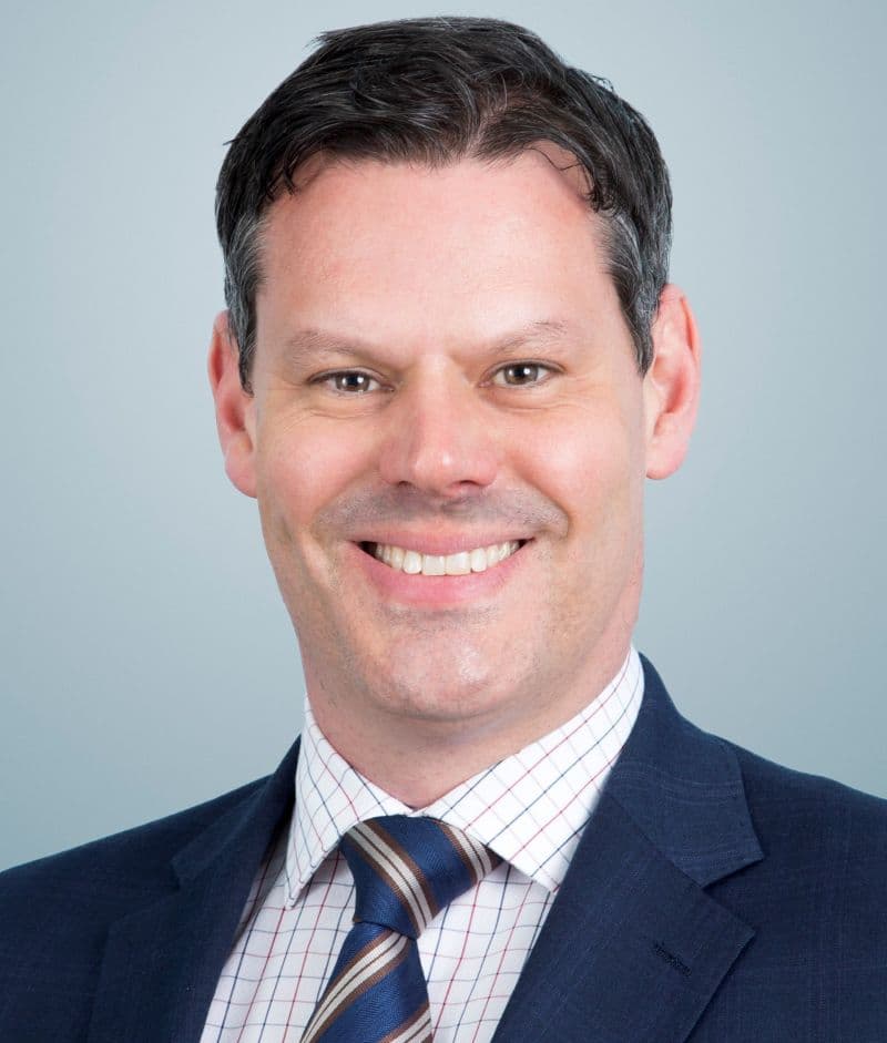 Professional headshot of a smiling middle-aged man with dark hair wearing a navy suit, striped tie, and checkered shirt against a gray background.