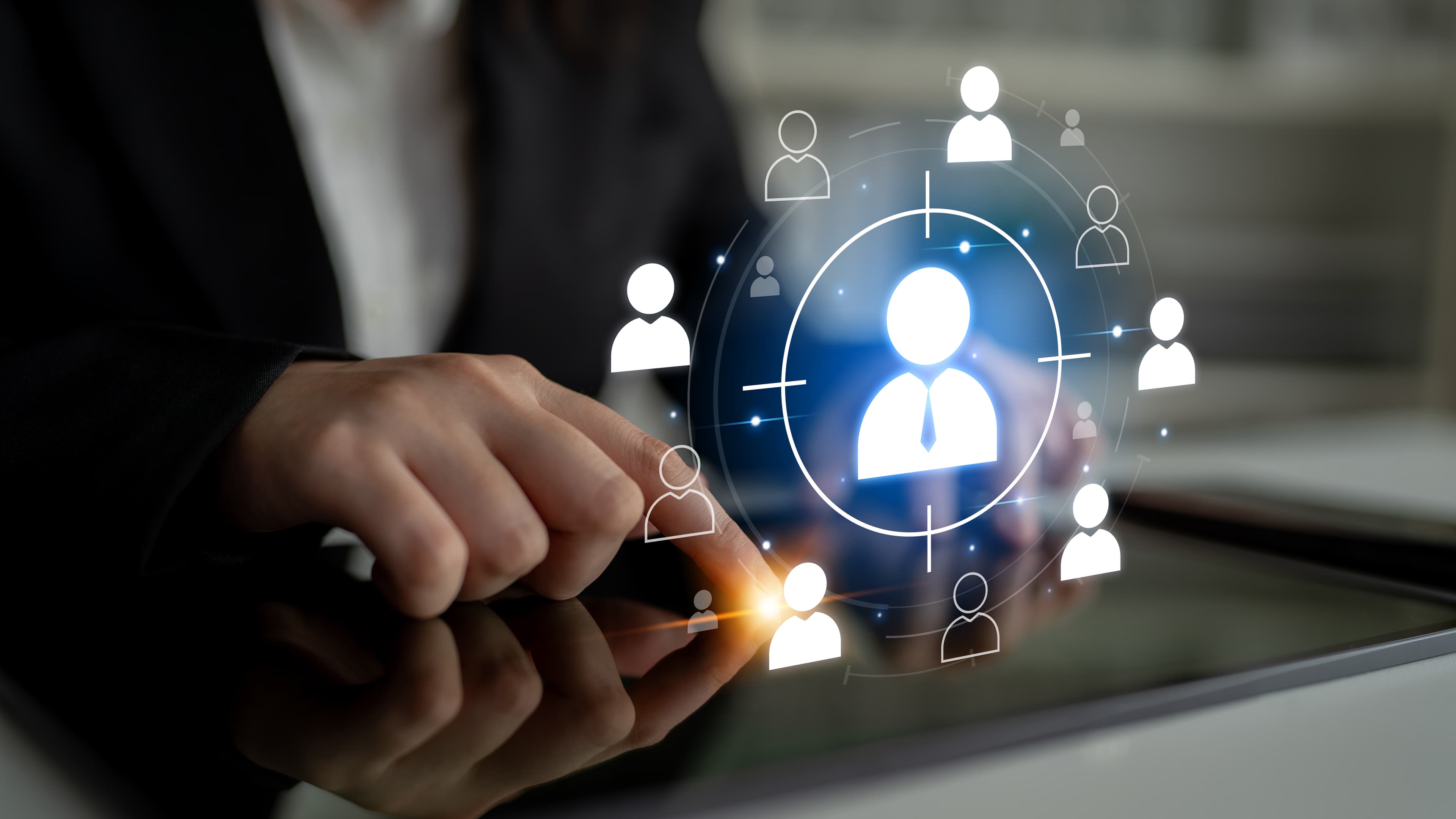 Person in a suit touching a tablet, with digital icons of people connected around a central figure, symbolizing network or recruitment.