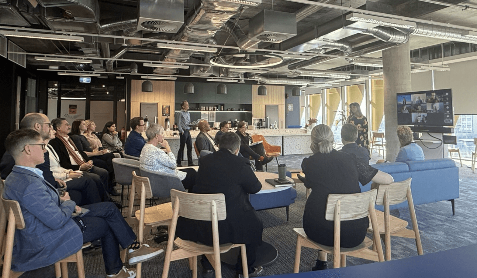 Group of professionals attending a business meeting in a modern office space with a presenter speaking and remote participants shown on a screen.