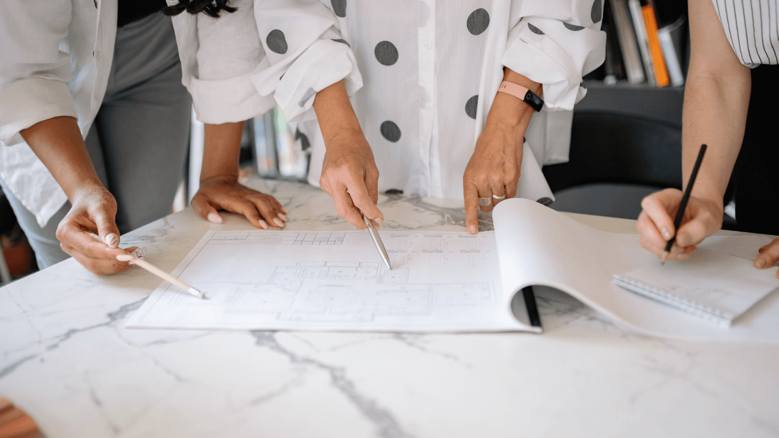 Three people reviewing architectural blueprints on a marble table, with one pointing using a stylus while another takes notes.