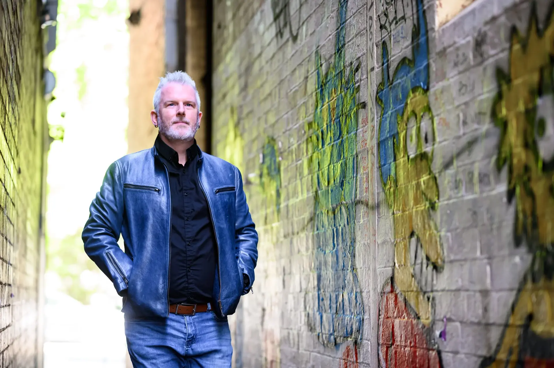 Middle-aged man with gray hair and beard wearing a blue leather jacket and jeans, standing in an urban alley with graffiti-covered brick walls.