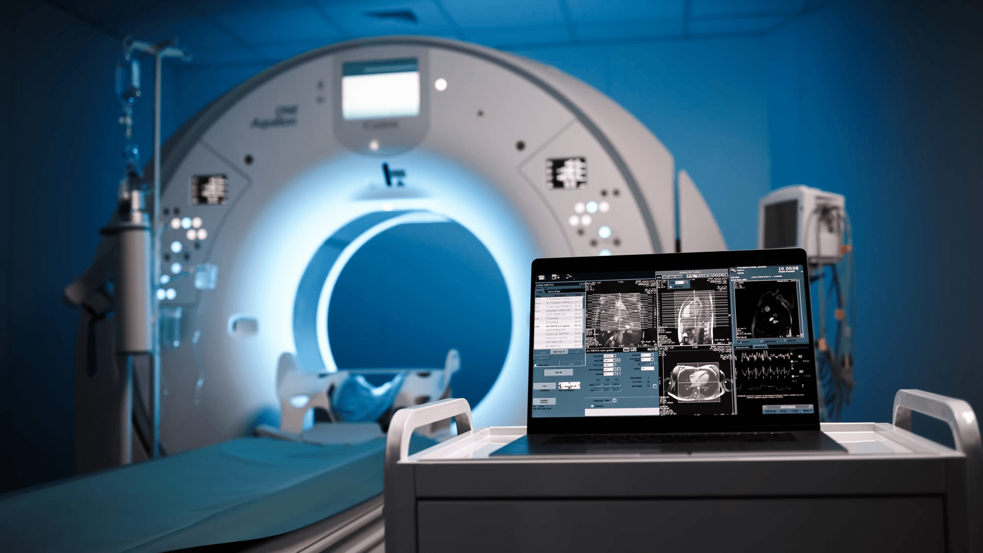 MRI machine in a dimly lit room with a laptop displaying scan images and data on a nearby cart.