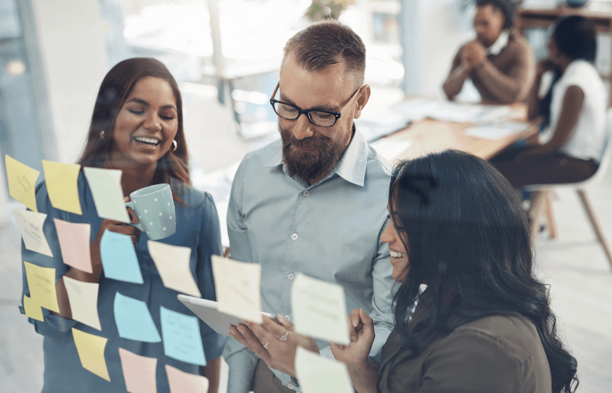 Three colleagues smile while discussing ideas in front of a glass wall covered with colorful sticky notes, in a bright office setting.