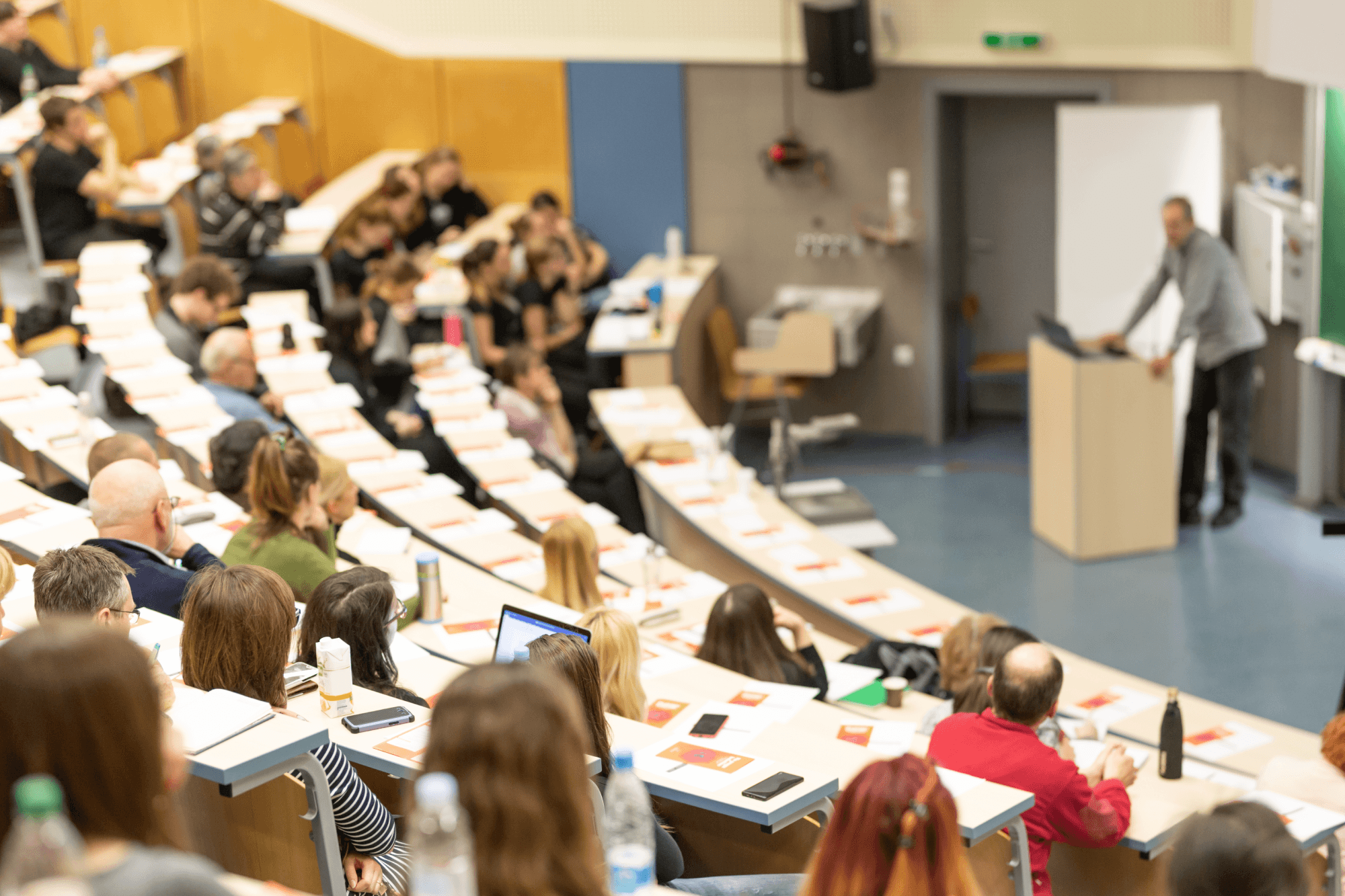 Lecture hall with students seated in rows, listening to a speaker at a podium. Desks are lined with papers and notebooks.