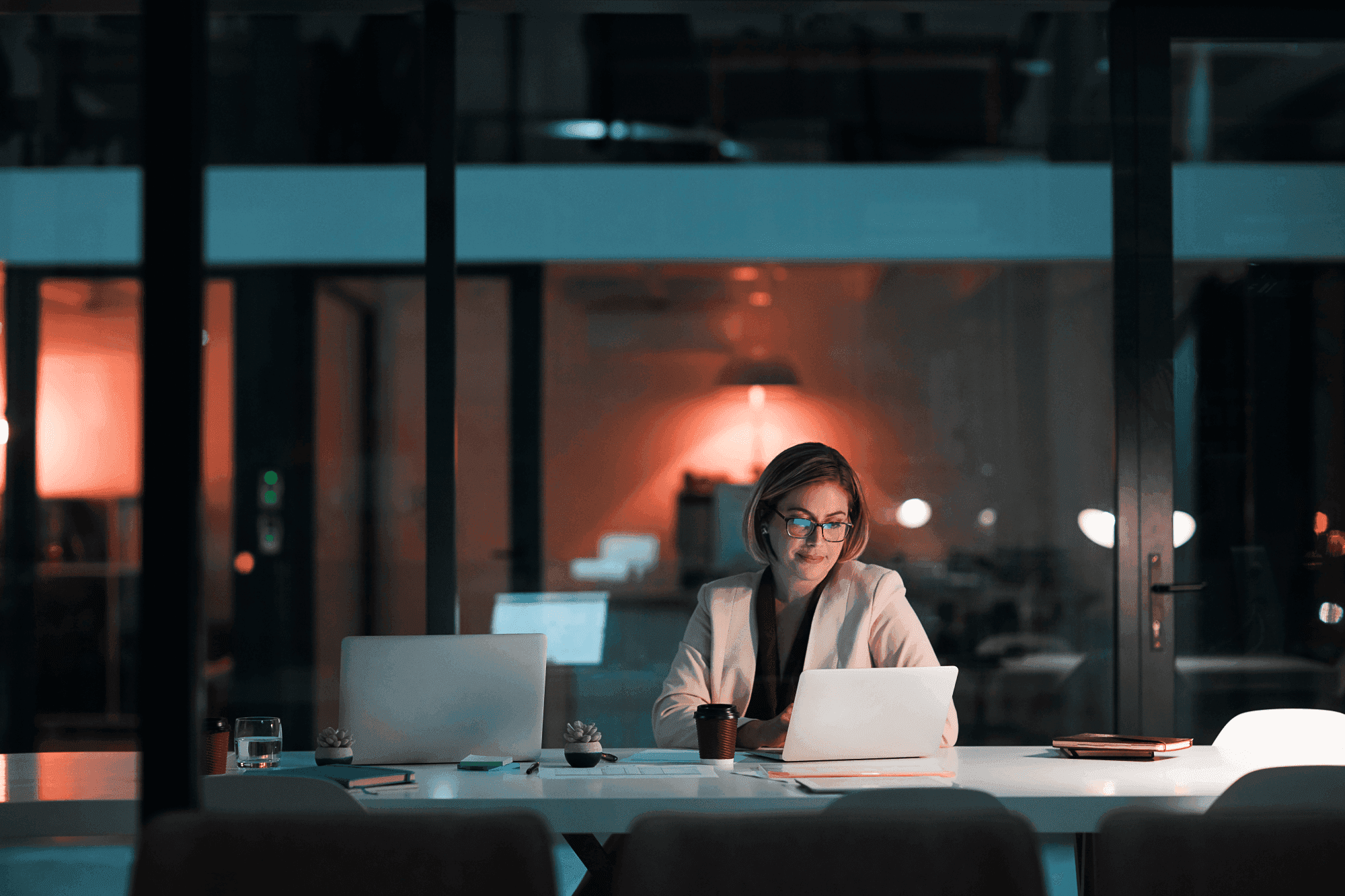 Woman in glasses working late at a dimly lit office desk with a laptop, papers, and a coffee cup.