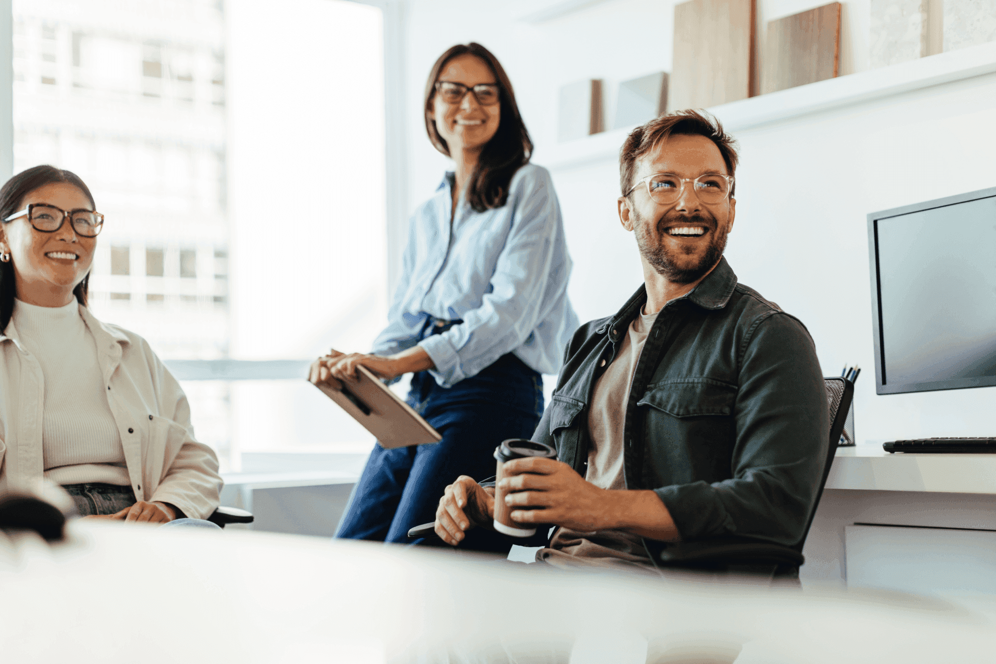 Three people in a bright office, smiling and engaging, one holding a clipboard, another with a coffee cup. A computer screen is visible.