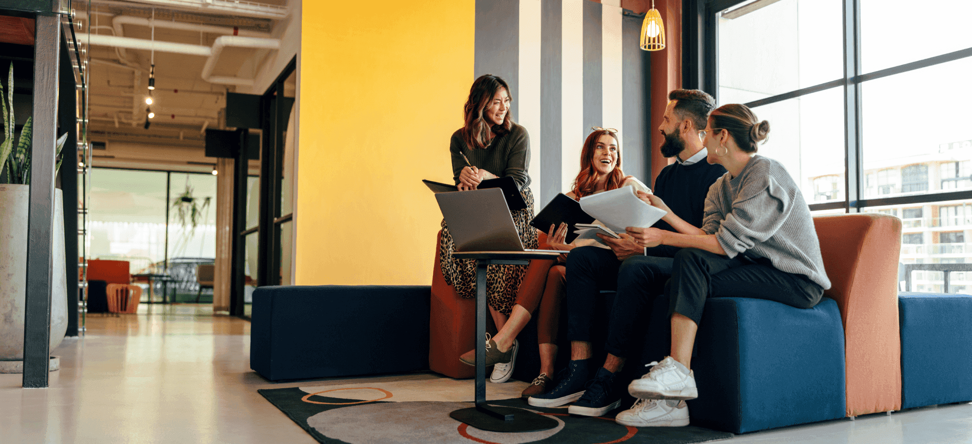 Four people casually discuss and smile in a modern office lounge, with a laptop and papers on their laps.