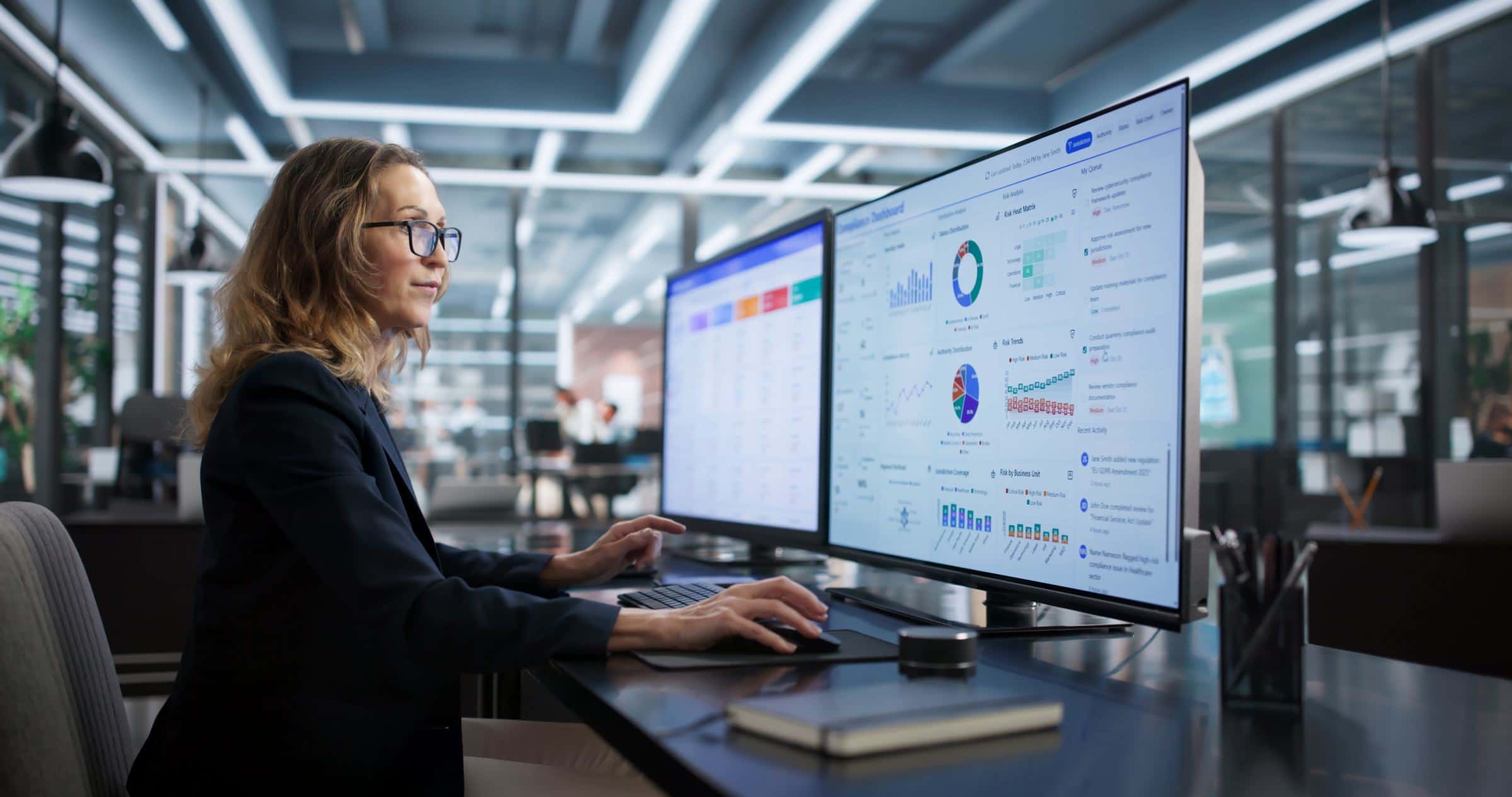 A woman at a desk in a modern office, focused on data visualizations displayed on two large computer monitors.