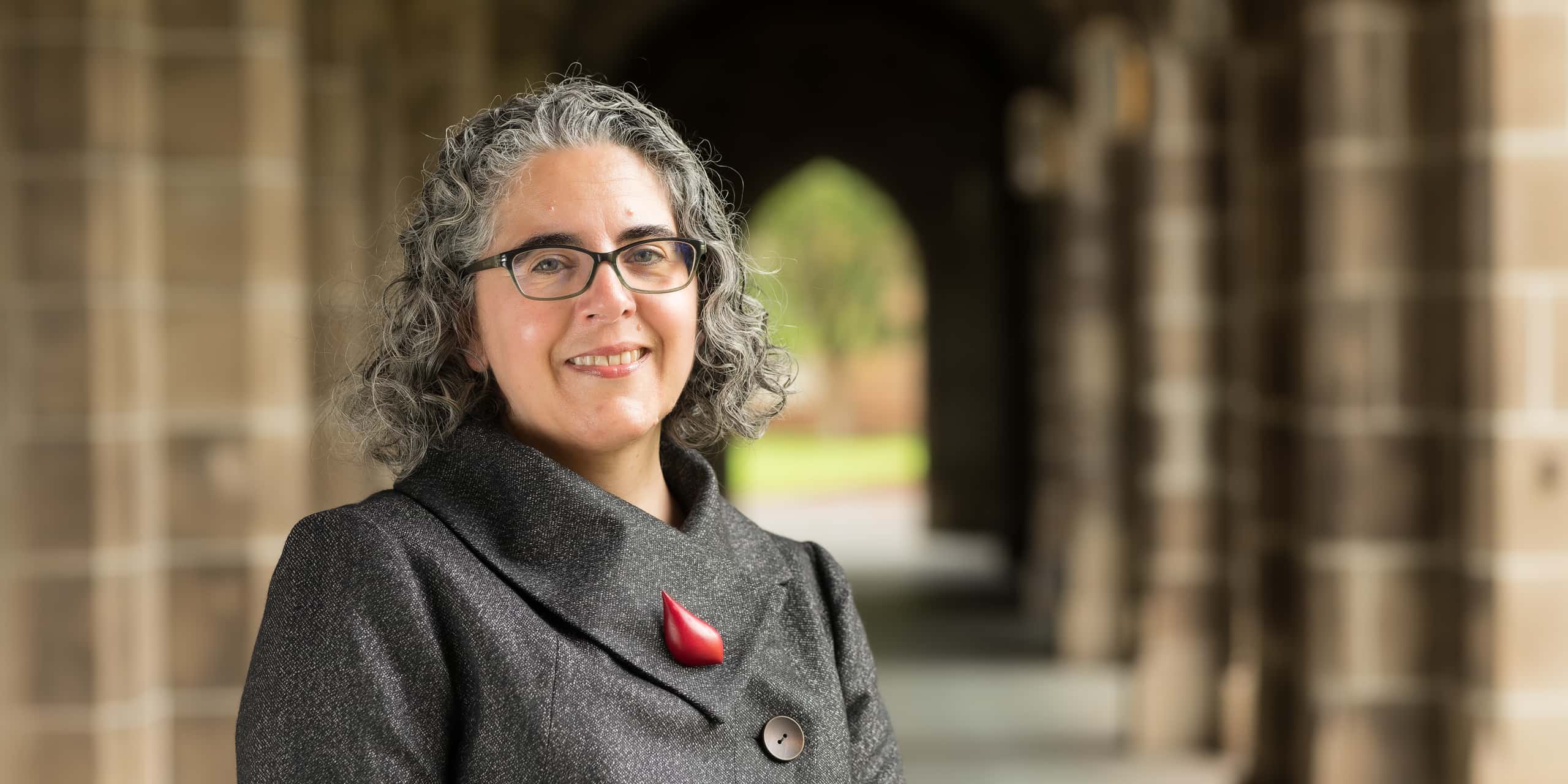 Woman with curly gray hair and glasses smiling in a stone archway corridor, wearing a gray coat with a red brooch.