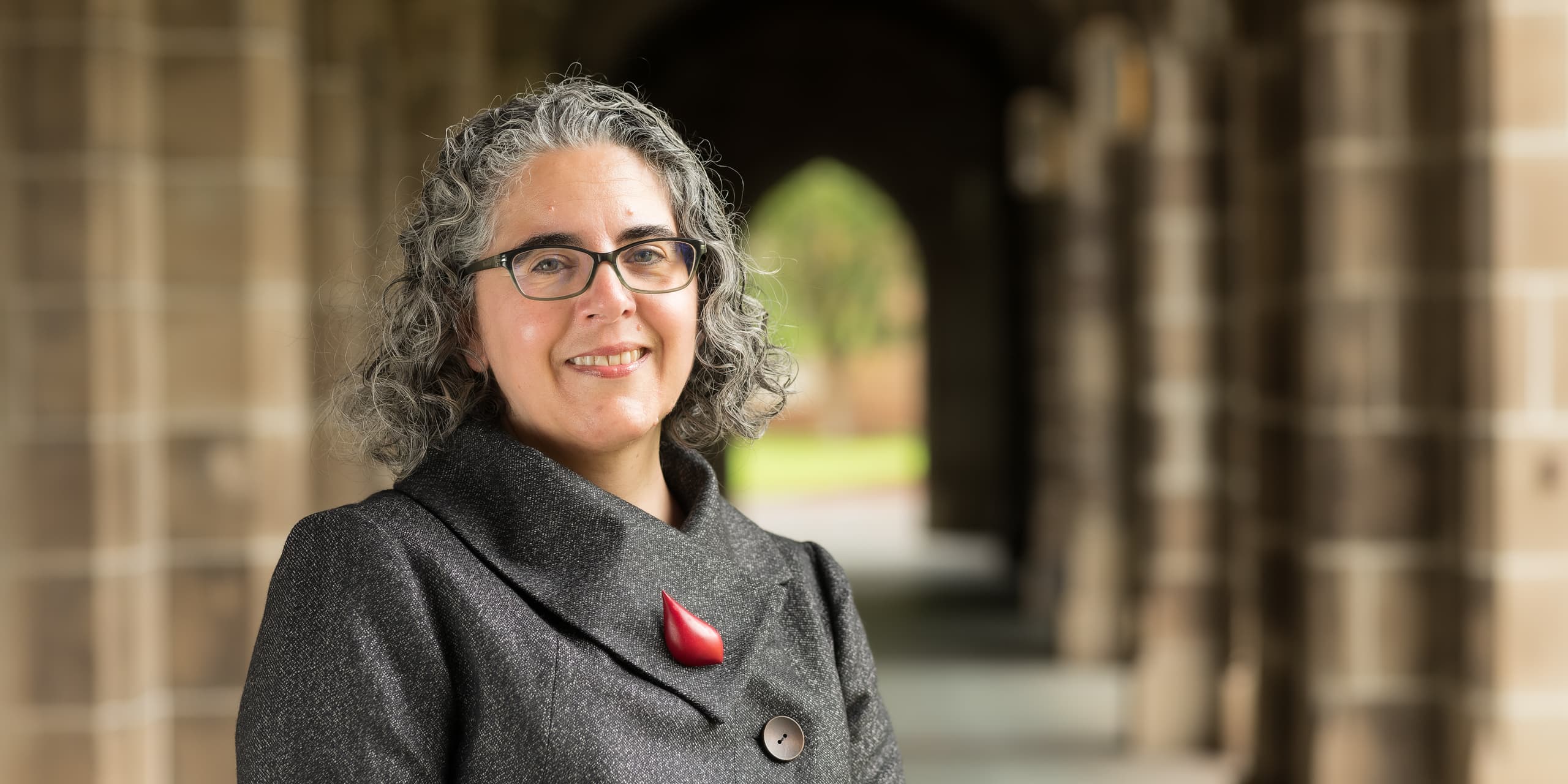 Woman with curly gray hair and glasses smiling in a stone archway corridor, wearing a gray coat with a red brooch.