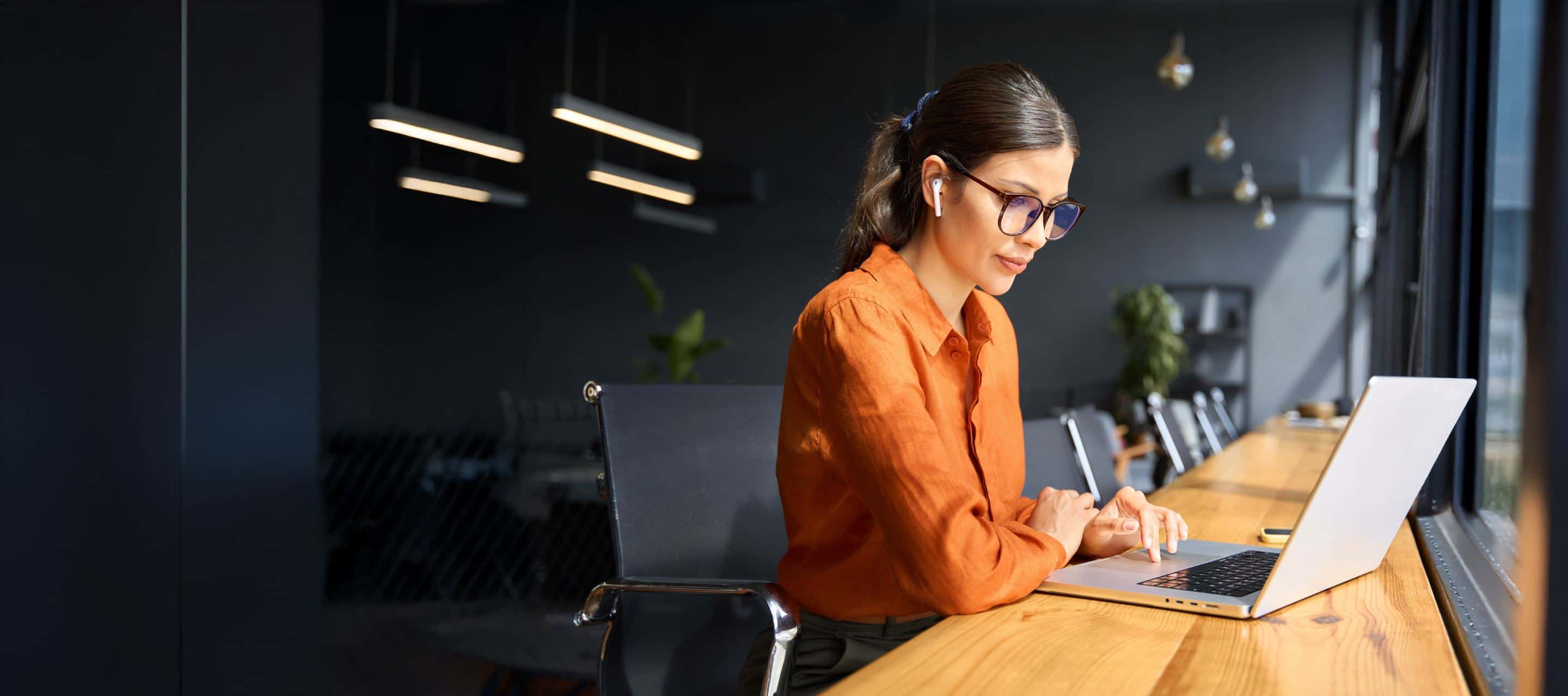 Woman in orange shirt and glasses wearing earbuds working on a laptop at a wooden desk in a modern dark office.