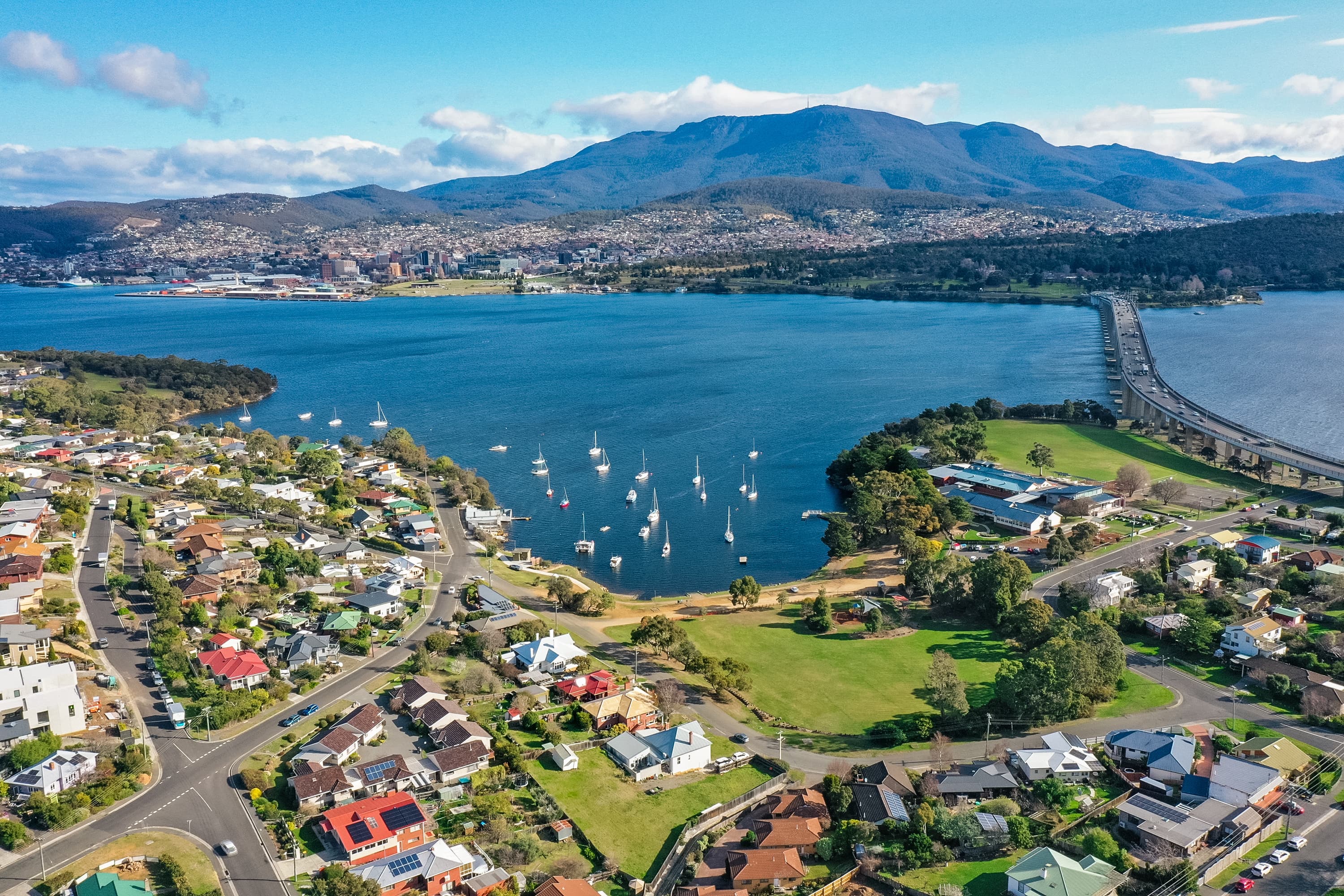 Aerial view of Hobart, Tasmania, showing the Derwent River, sailboats, residential suburbs, a bridge, and Mount Wellington in the background.