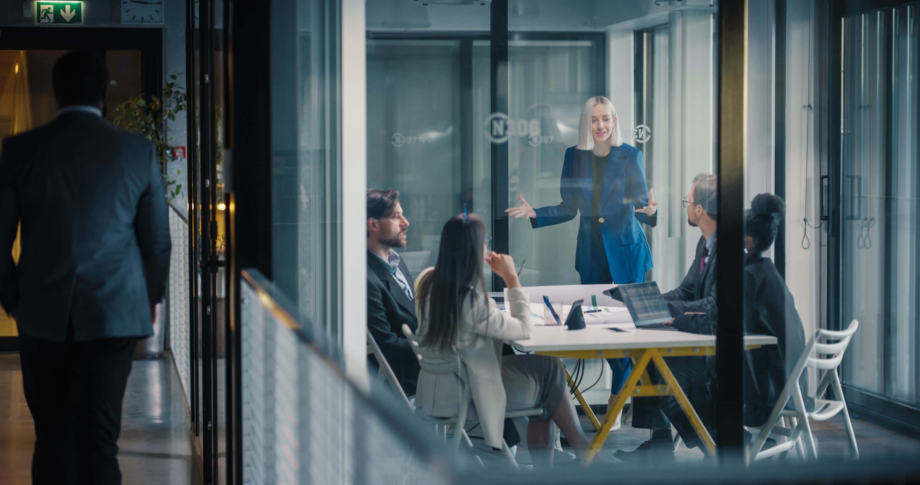 Woman in blue suit presenting to colleagues in a modern glass-walled conference room during a business meeting.
