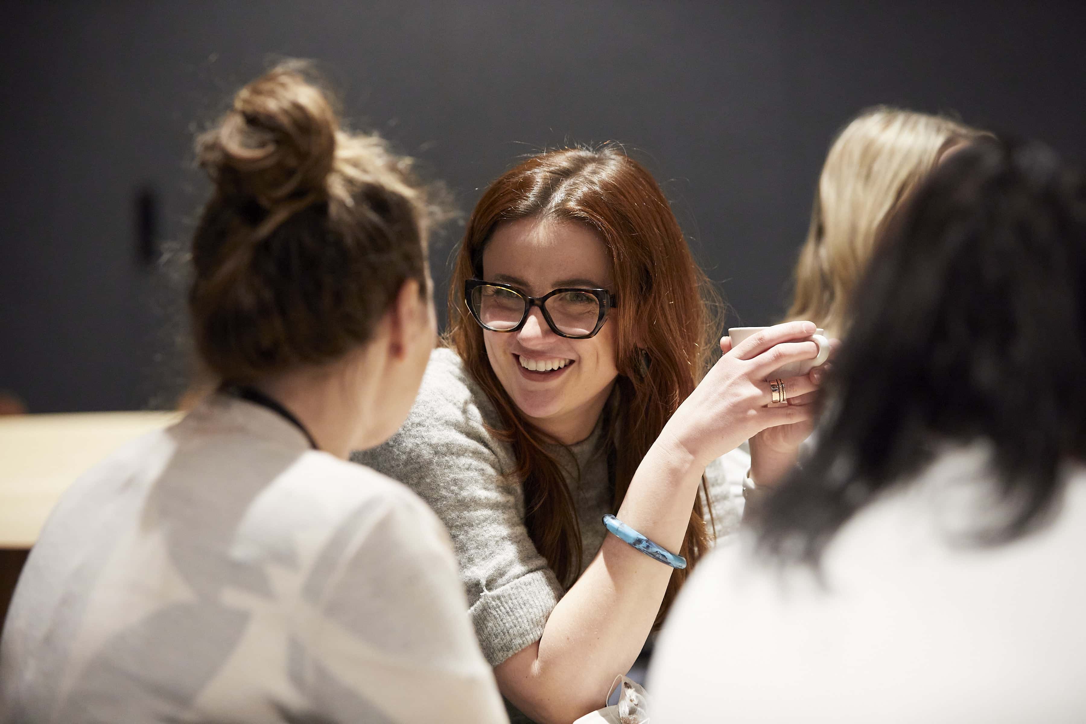Smiling woman wearing glasses holds a cup while chatting with friends around a table.