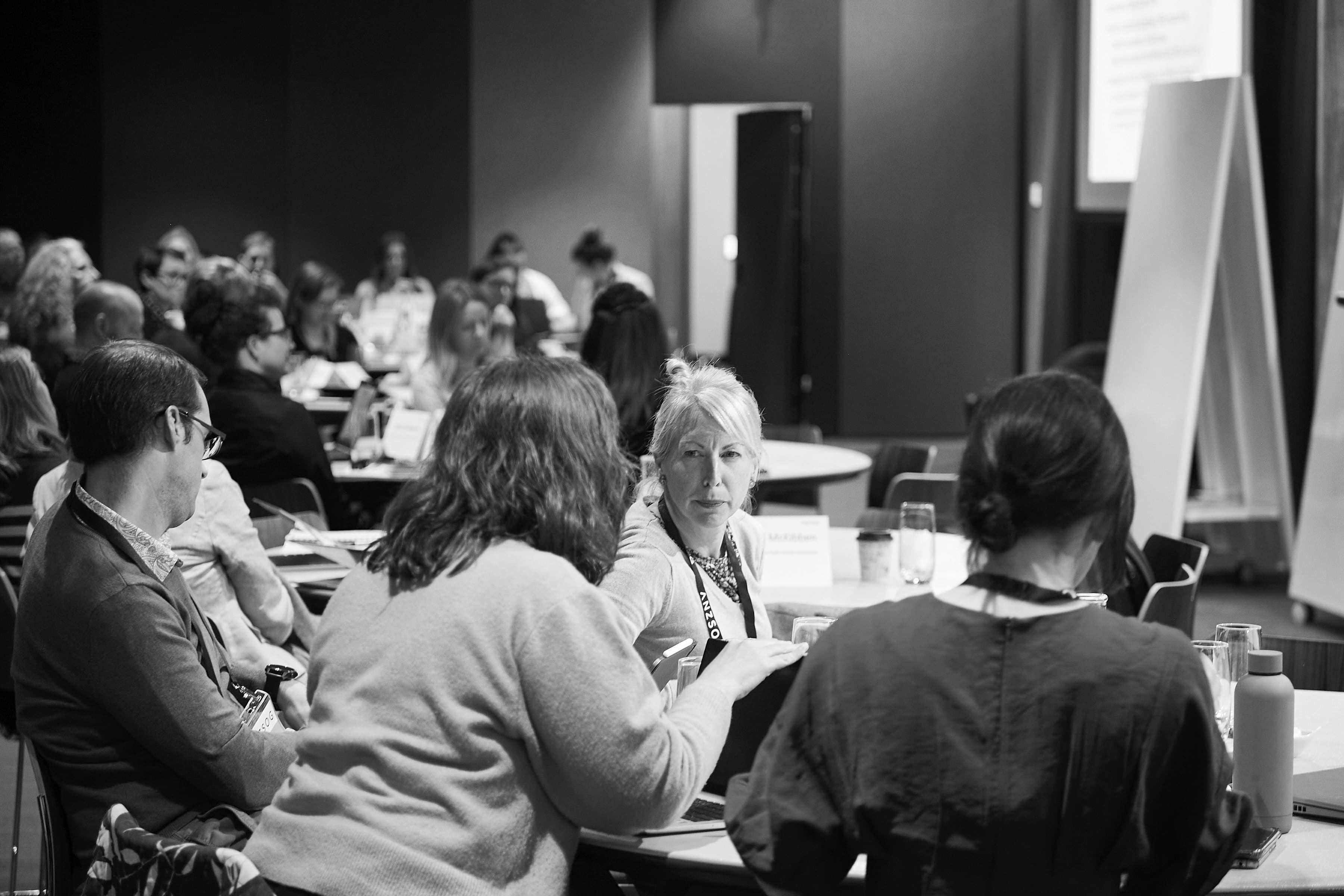Black-and-white photo of a workshop: a light-haired woman looks intently across a table while colleagues talk and take notes.