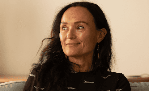A woman with long dark hair and gold earrings smiles while looking to the side, sitting on a couch. She wears a dark top with a subtle pattern.