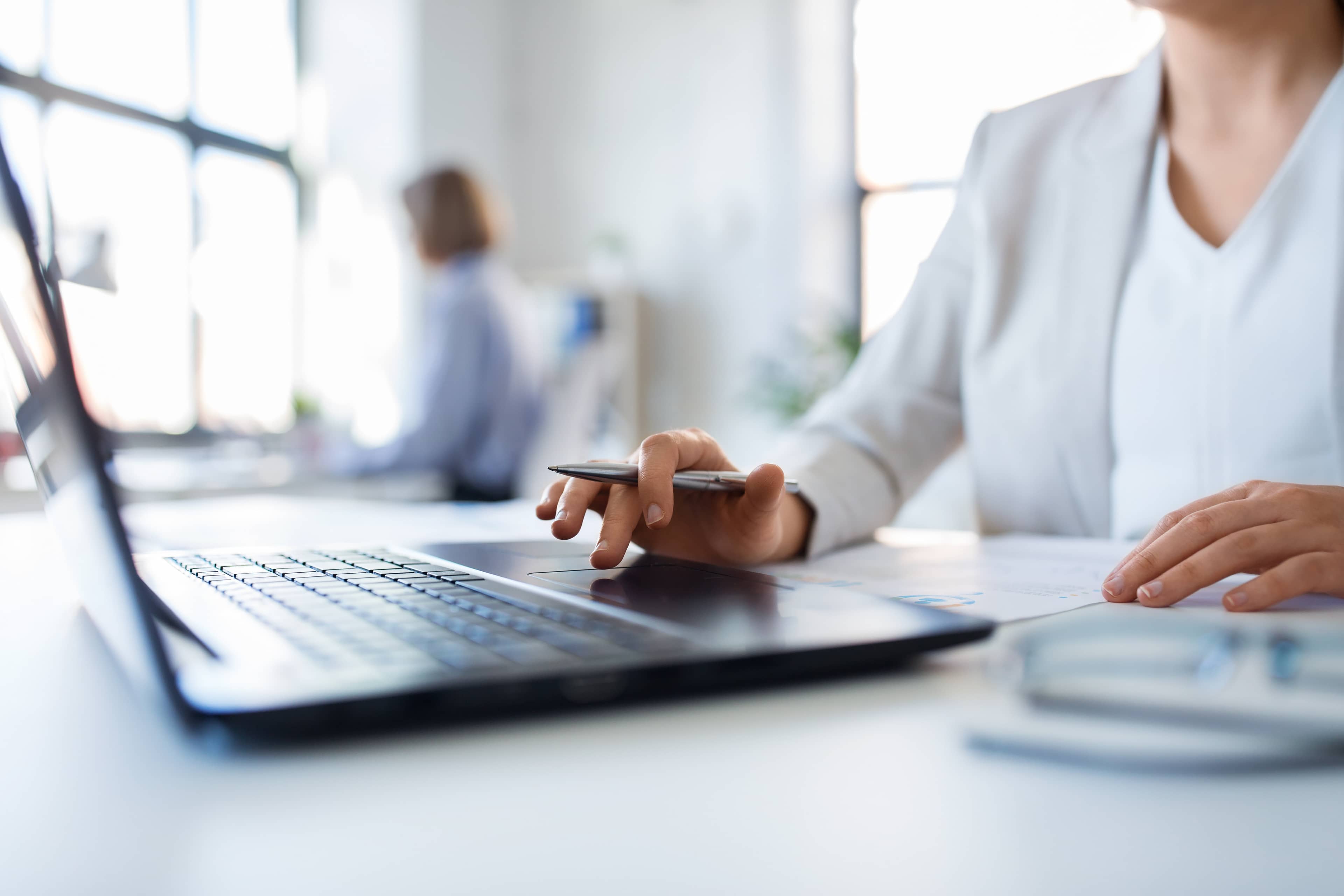 A person in a white outfit works on a laptop, holding a pen, in a bright office with large windows. Another person is blurred in the background.