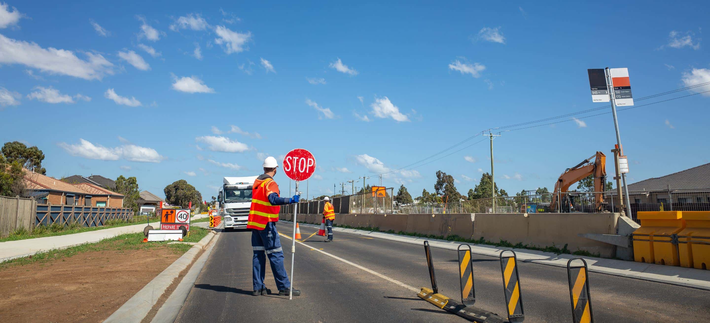 Road worker in orange vest and white helmet holding a stop sign at a suburban construction zone with barriers and excavator nearby.