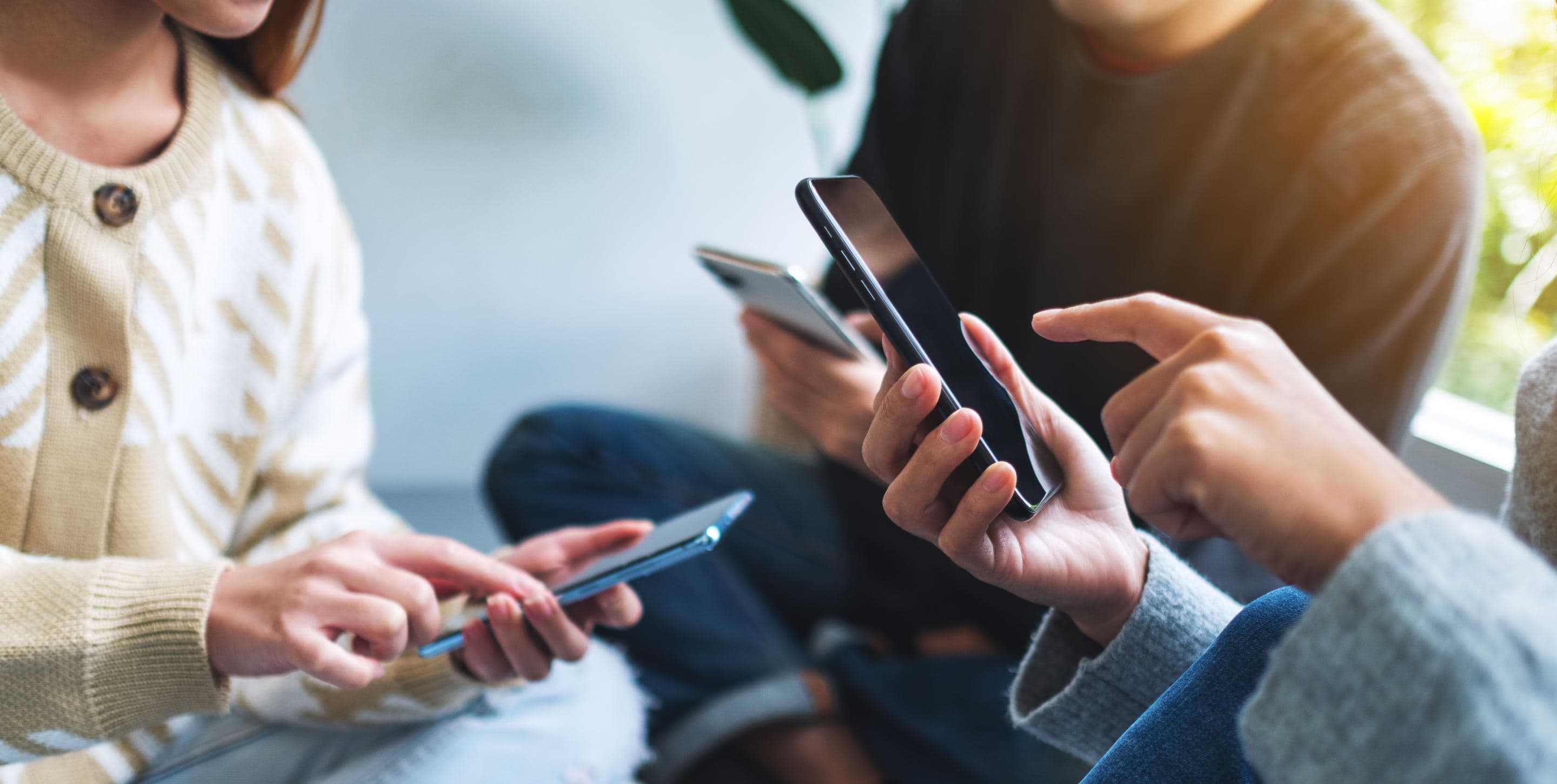 Three people sitting together, each using and scrolling on their smartphones, close-up view of hands and devices.
