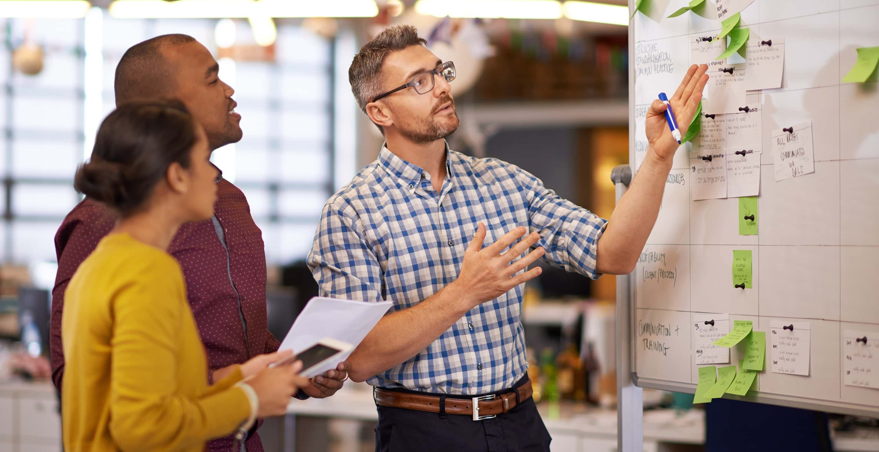 Three colleagues in a office discussing a whiteboard covered with sticky notes and written tasks during a team planning meeting.