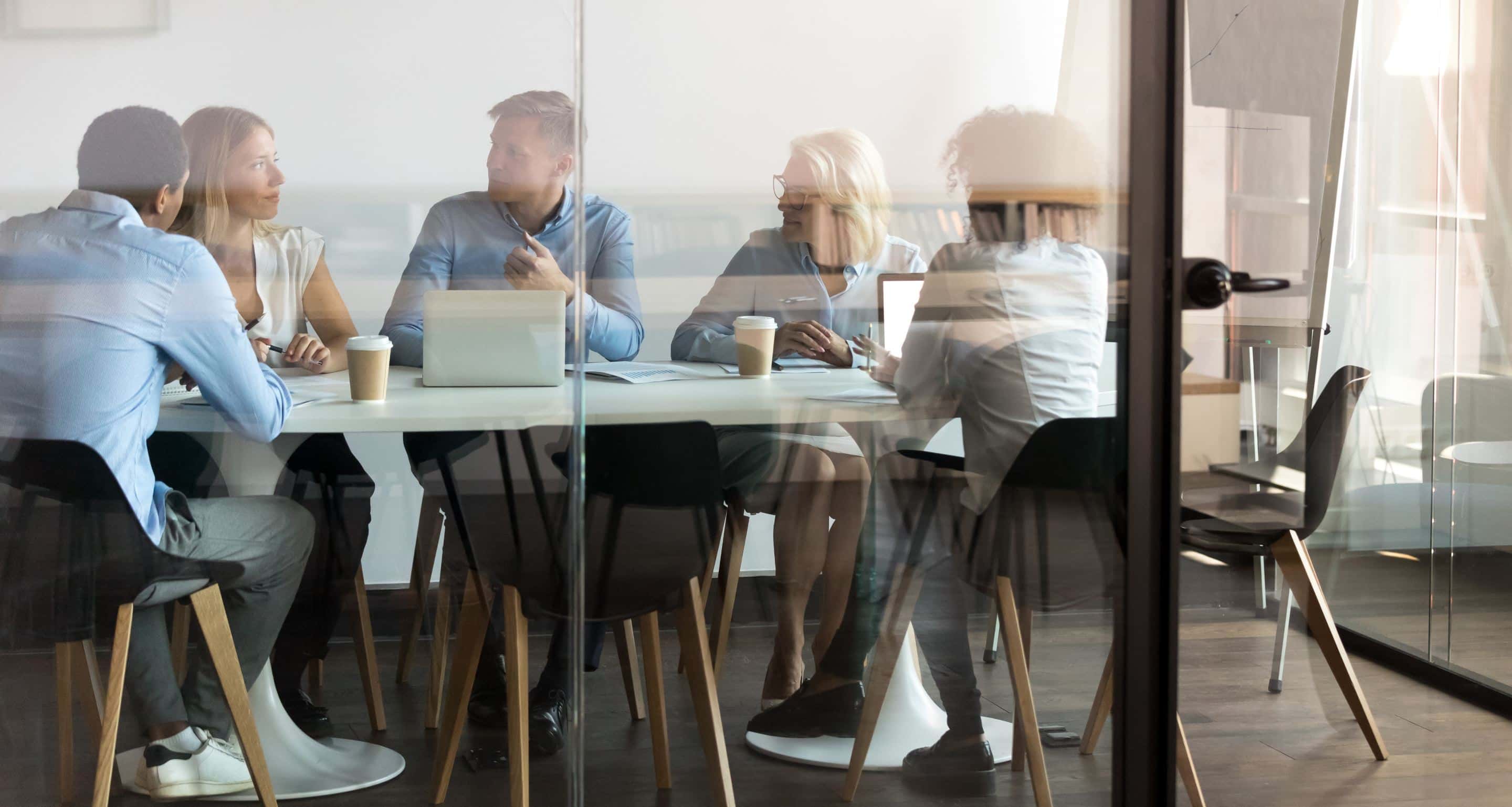 Five diverse professionals having a business meeting in a glass-walled conference room with laptops and coffee cups on the table.