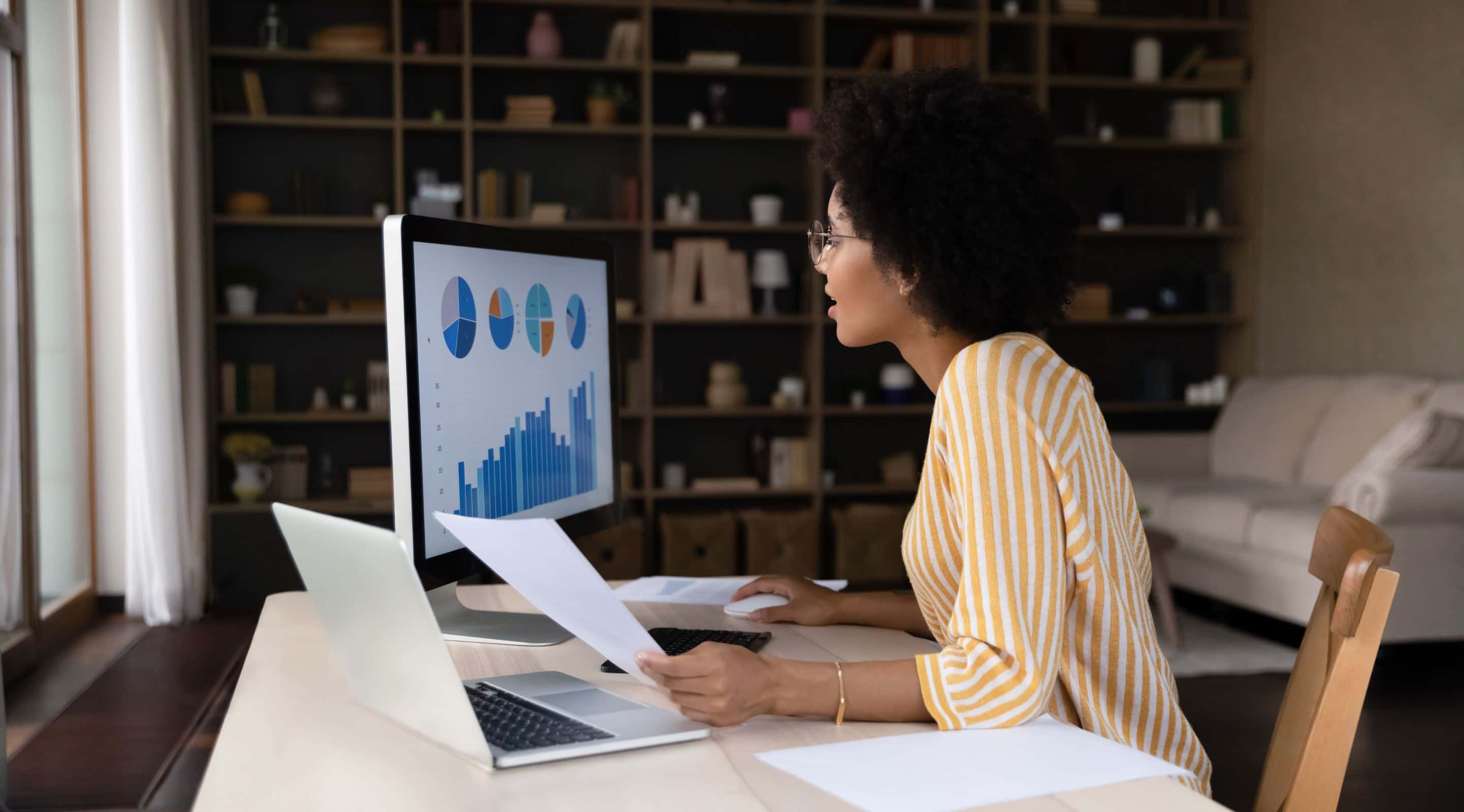 Woman in yellow striped shirt reviewing data charts on desktop monitor while holding documents at home office desk with bookshelf background