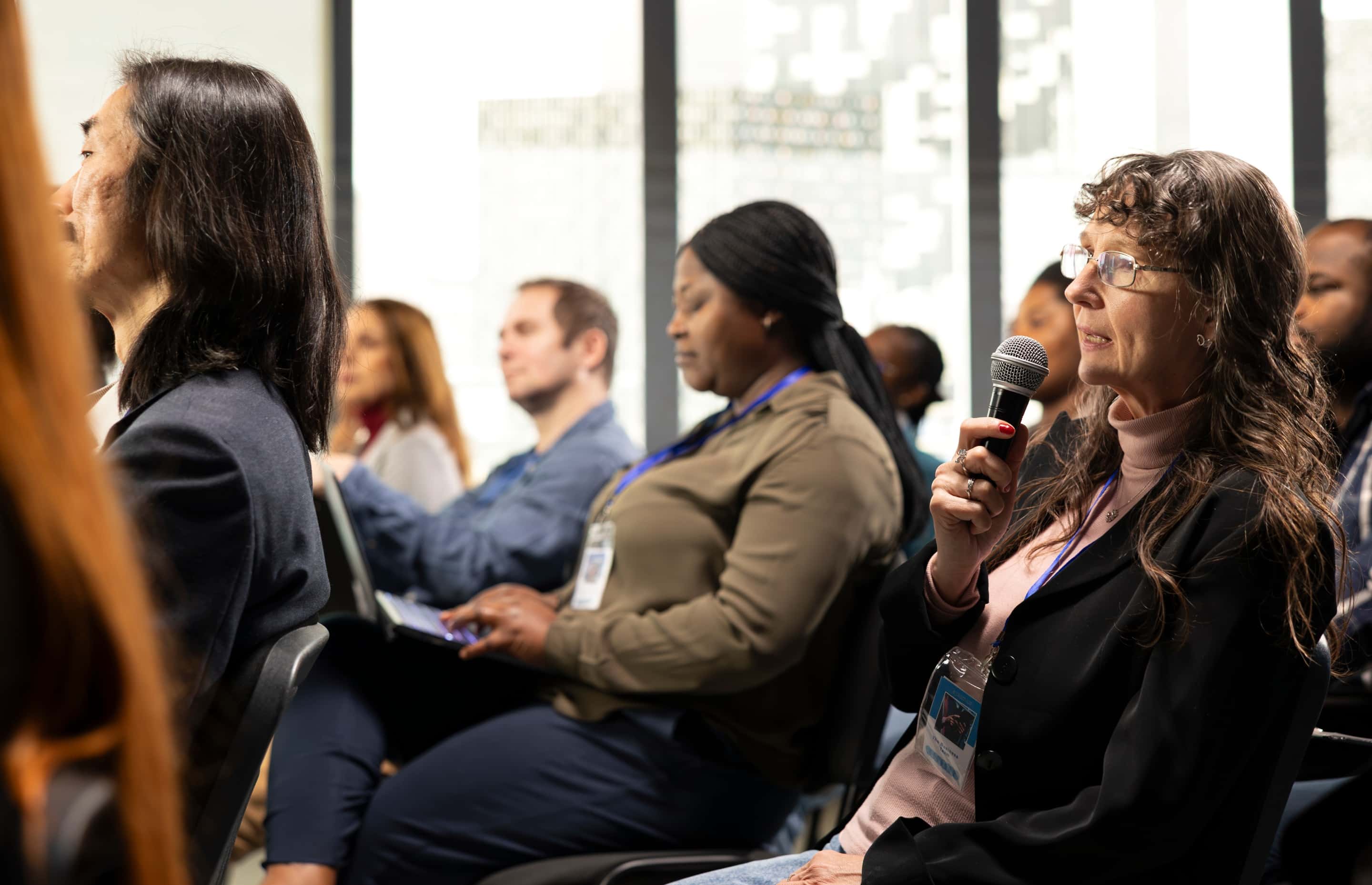 A woman with glasses holds a microphone at a conference while diverse attendees with lanyards sit in rows behind her.