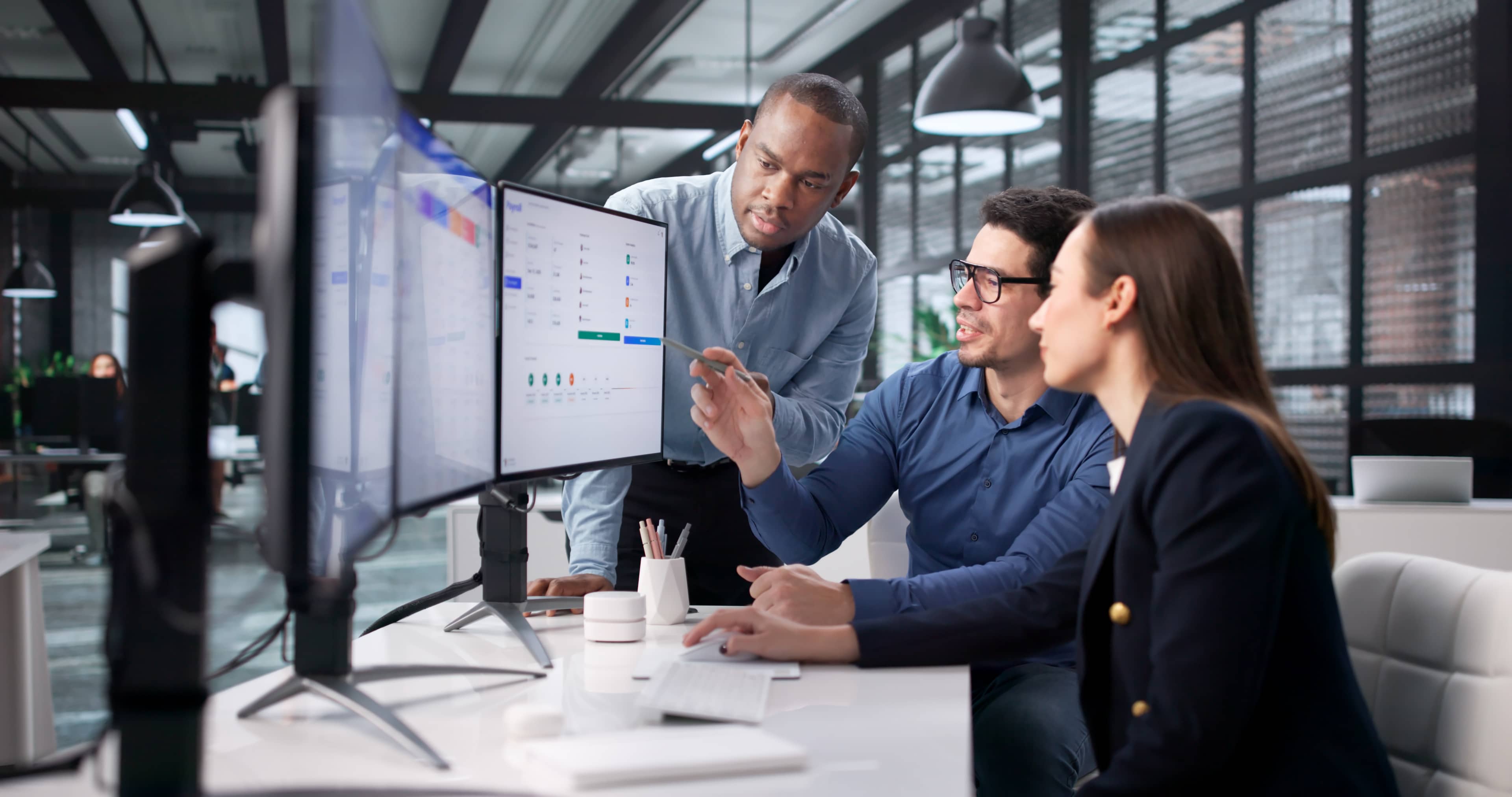 Three professionals collaborate at a desk, focusing on dual monitors displaying charts in a modern office setting.