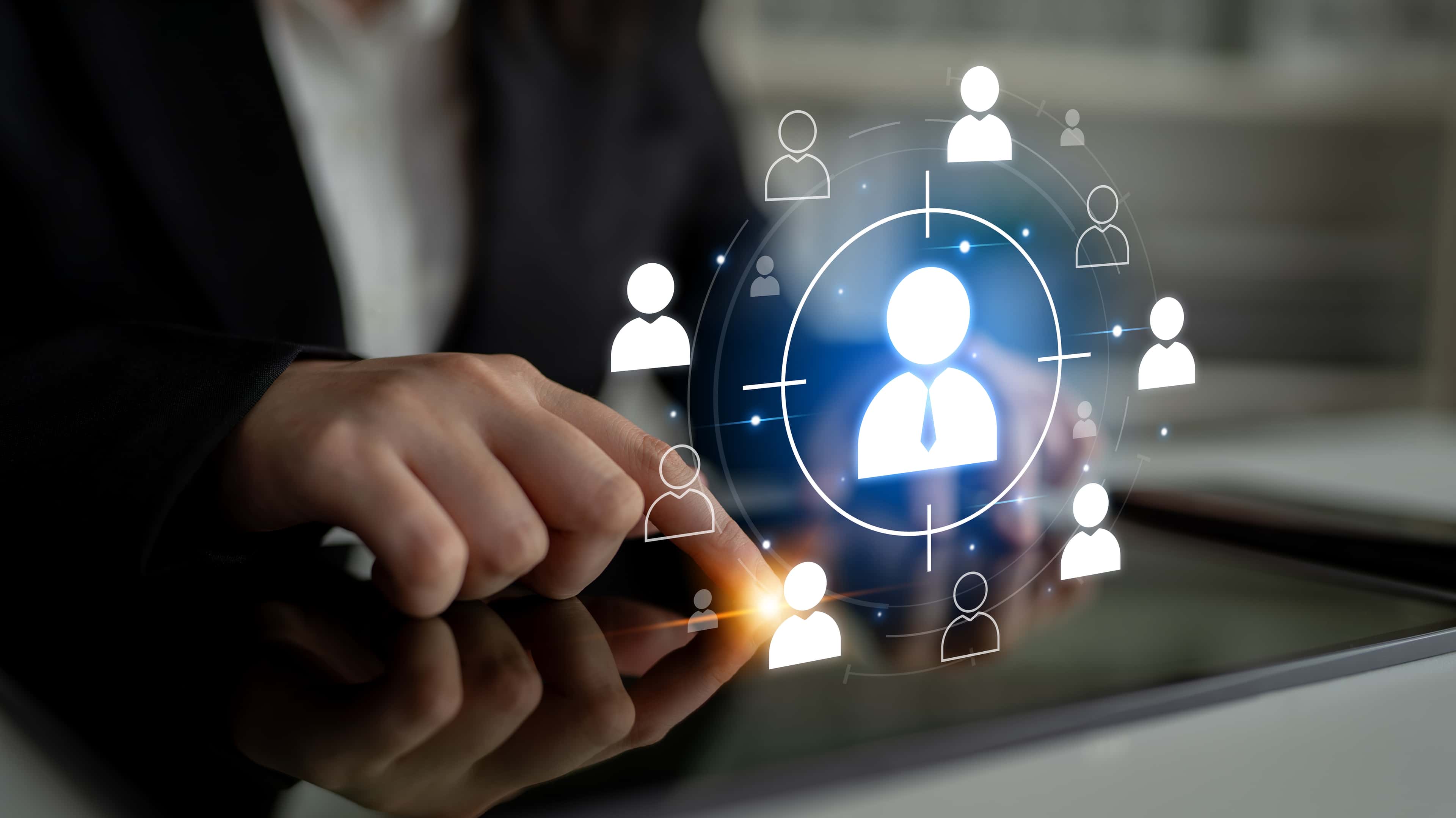 Person in a suit touching a tablet, with digital icons of people connected around a central figure, symbolizing network or recruitment.