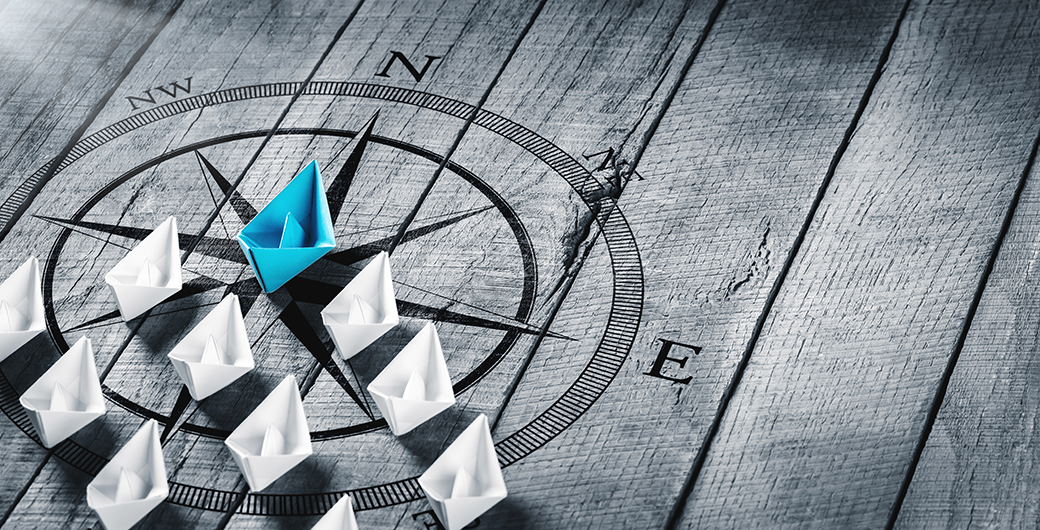 Blue Paper Boat Leading A Fleet Of Small White Boats With Compass Icon On Wooden Table With Sunlight